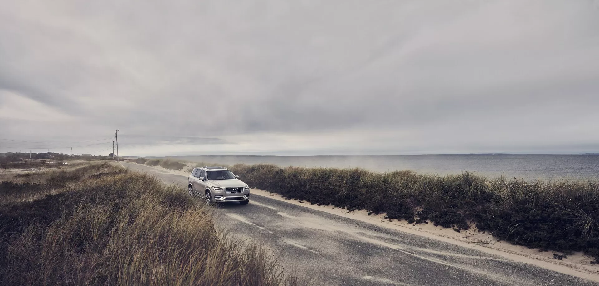 Širokouhlá fotografia Volvo XC90 z roku 2015 (2. generácia) jazdiaceho po úzkej pobrežnej ceste. Auto sa približuje ku kamere, zvýrazňujúc prednú mriežku, svetlomety a nárazník. Na pozadí so zamračenou oblohou a oceánom vyniká svetlá kovová farba SUV. Táto scéna zdôrazňuje kombináciu luxusu a dobrodružstva vozidla, naznačujúc pokročilé technologické a bezpečnostné funkcie pre jazdné podmienky na pobreží.