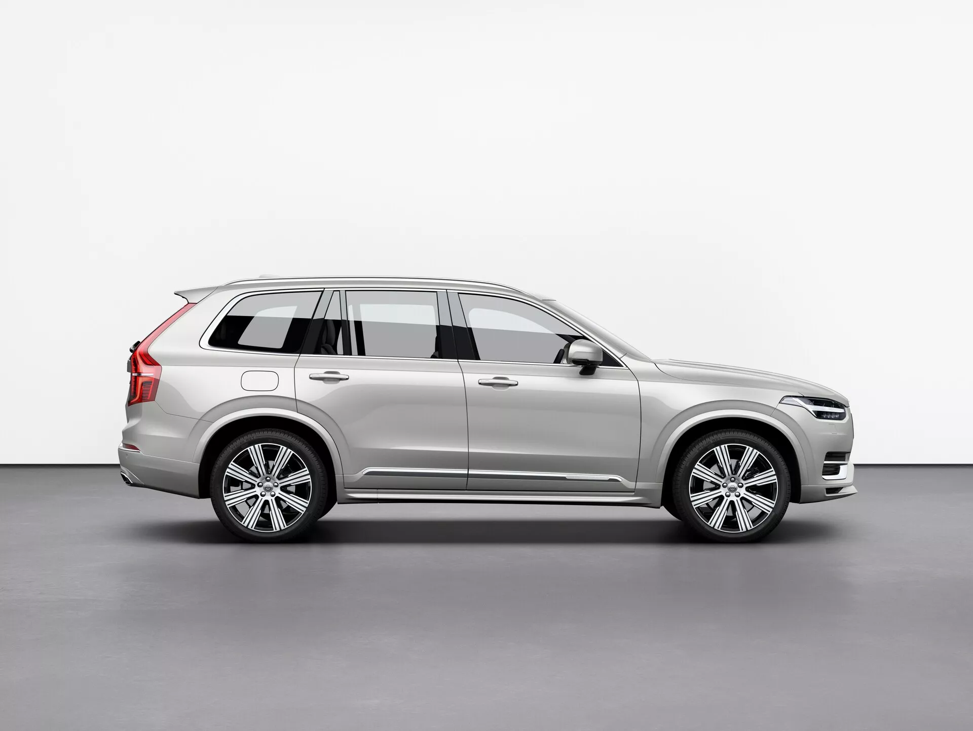 Bočný profil strieborného SUV Volvo XC90 2015 (2. generácia), zobrazujúci jeho hliníkové kolesá, strešné nosiče, bočné zrkadlo s integrovaným smerovkou a detailné línie karosérie, umiestnený centrálne na jednoduchom pozadí.
