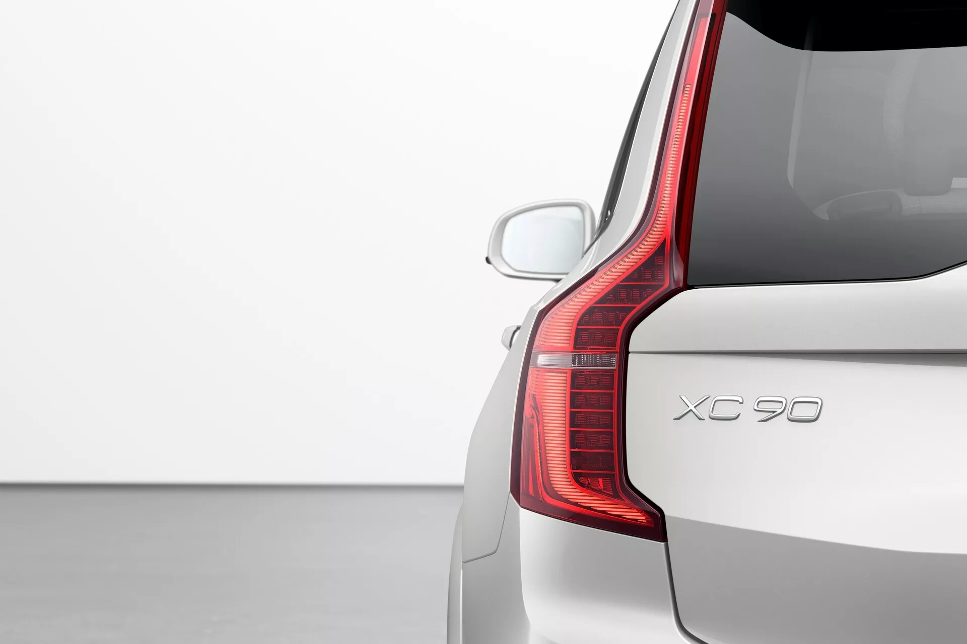 Detailný pohľad na zadnú časť strane spolujazdca modelu Volvo XC90 z roku 2015 (2. generácia), ktorý ukazuje charakteristický dizajn LED zadných svetiel, zadné veko a zadný nárazník. Uhol zozadu pod tromi štvrtinami zdôrazňuje vertikálne rozšírenie zadného svetla a jeho jedinečný tvar, ktorý sa zakrivuje dopredu do strany. Obrázok zvýrazňuje aj zadné čelné sklo s aerodynamickým strešným spojlerom, viditeľný zadný parkovací senzor a jednofarebný lak v jednoduchom štúdiovom prostredí, so zameraním na dizajn a vlastnosti auta.