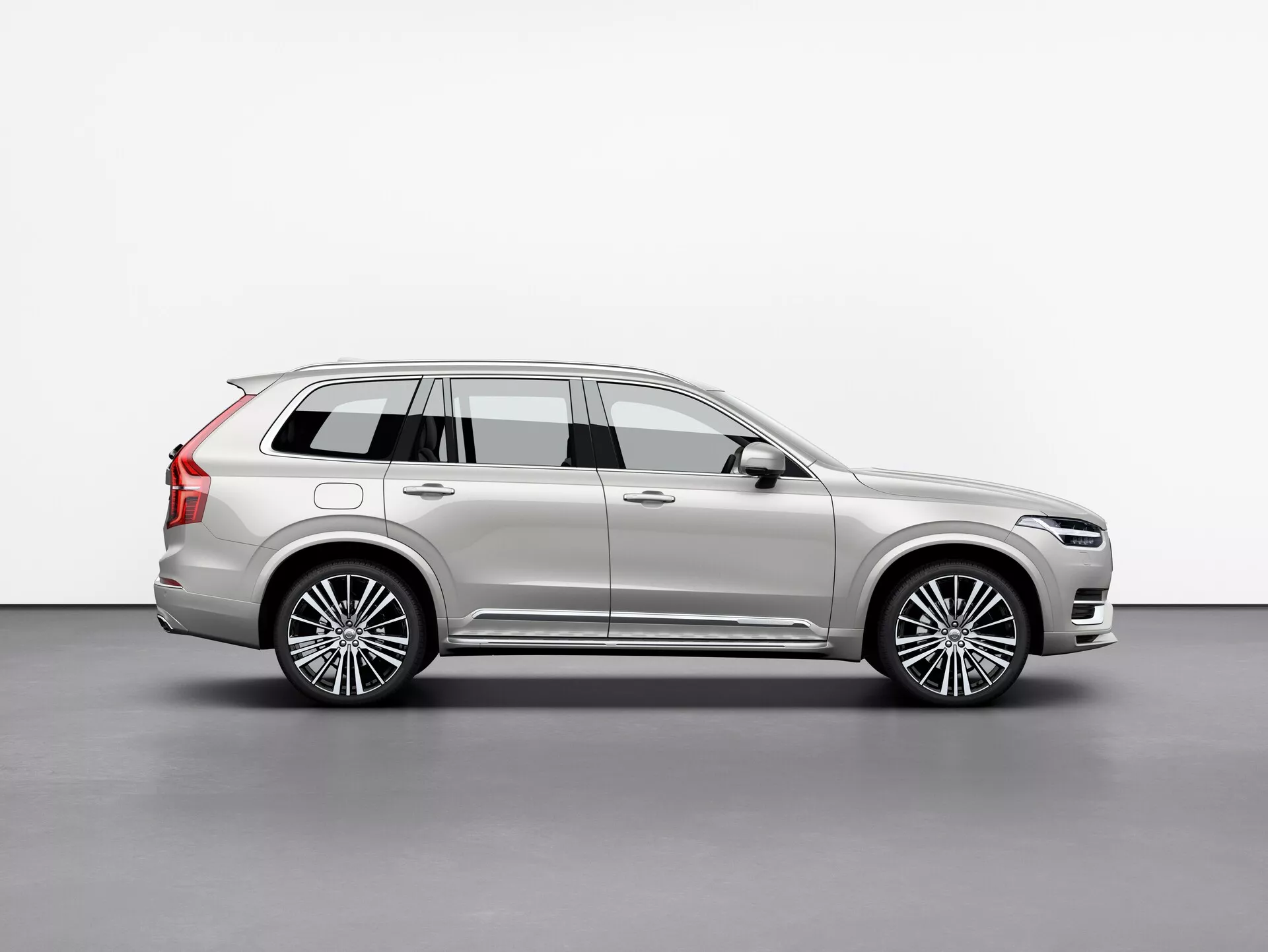 Bočný profil vozidla Volvo XC90 2015, luxusného SUV v plnej veľkosti, zachytený v štúdiu na bielom pozadí. Obrázok zobrazuje dlhý rázvor vozidla, štyri dvere, zadné dvere, viacpaprskové zliatinové kolesá, chrómové ozdoby, metalický lak a výrazné dizajnové prvky, ako sú bočné zrkadlá a strešné nosiče. Starostlivo osvetlená scéna zvýrazňuje línie a krivky karosérie, odrážajúc jej prémiový charakter. Vozidlo je umiestnené tak, aby poskytovalo jasný pohľad na jeho celkovú dĺžku, výšku a postavu, čo naznačuje SUV s kapacitou pre tri rady cestujúcich a nákladu.