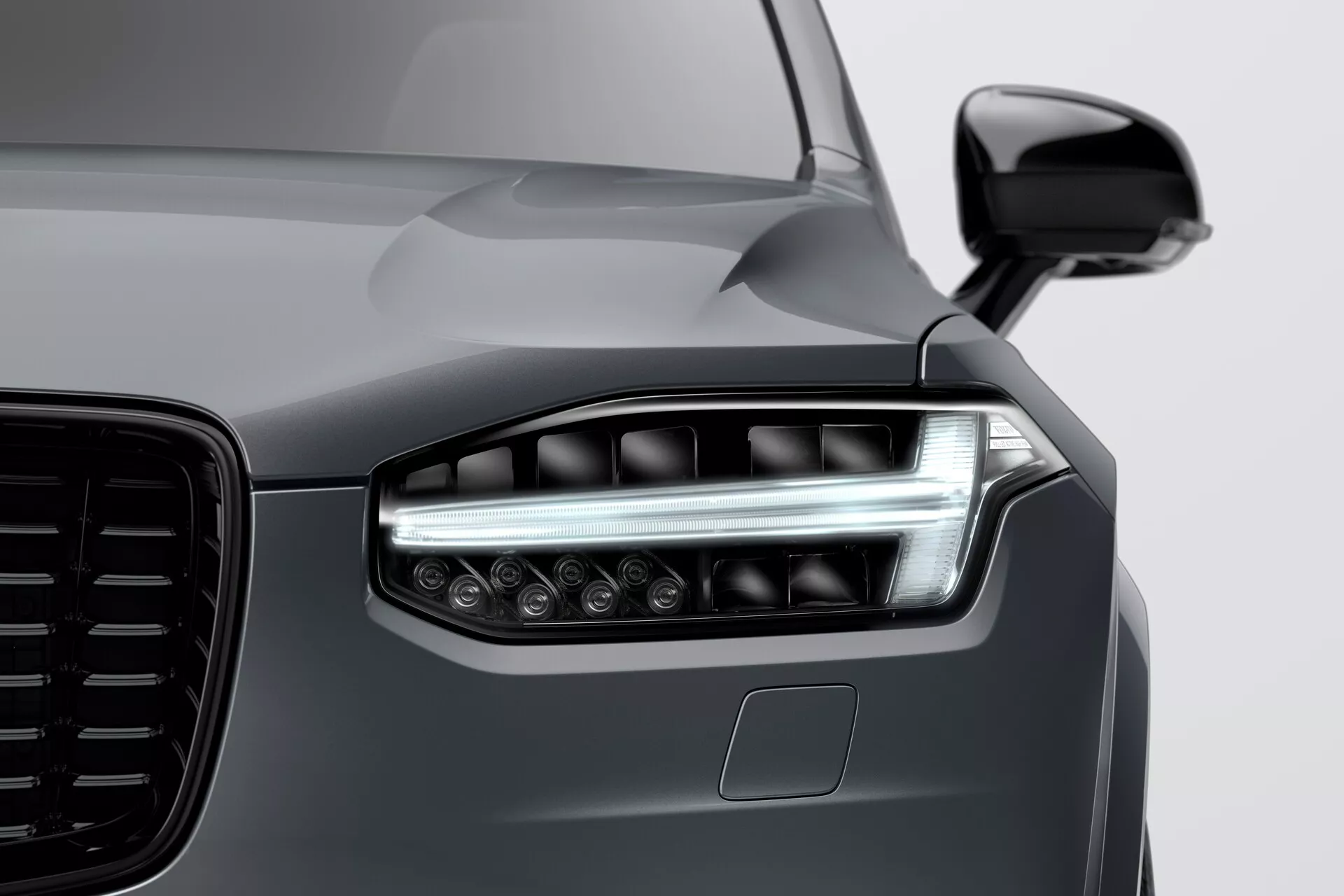 Detailný záber na prednú časť Volvo XC90 2015 (2. generácie), so zameraním na ľavé svetlo s modernou LED alebo laserovou technológiou, dvojitými svetelnými prvkami, elegantnými dennými bežnými svetlami a niekoľkými projekčnými šošovkami pre lepšiu viditeľnosť. Obrázok zdôrazňuje veľkú esteticky dizajnovanú mriežku, aerodynamické bočné zrkadlo vo farbe karosérie a možný senzor ADAS pod svetlom. Hladký, reflektívny povrch v hlbokej metalickej šedej farbe podtrhuje elegantné, ale svalnaté línie kapoty a blatníka, čo ukazuje na vysokokvalitnú farbu a špičkový dizajn vozidla.