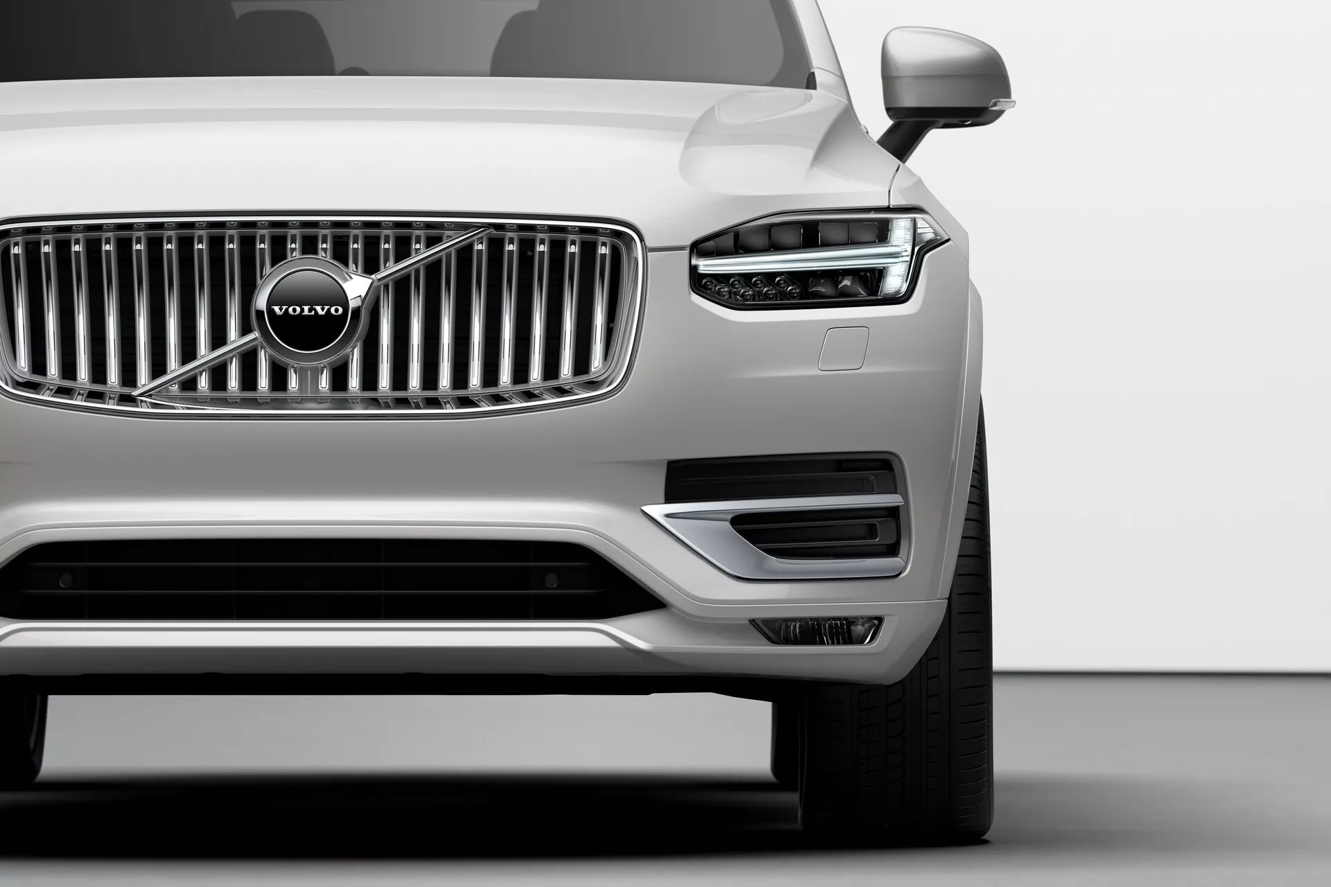 Nízky uhol záberu prednej ľavej štvrtiny strieborného Volvo XC90 (2. generácia) z roku 2015 v štúdiu, ktorý ukazuje charakteristický LED denný svetelný vzor v tvare Thorovho kladiva, vertikálne chrómované rebrované mriežky s emblémom Volvo, minimalistický predný nárazník s nasávacími otvormi, ľavé spätné zrkadlo, viacramenné zliatinové koleso a bezchybný metalický povrch, ilustrujúci pokročilú technológiu osvetlenia, dizajn značky a aerodynamické vlastnosti.