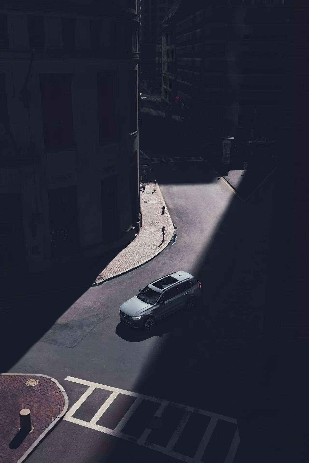 Letecký pohľad na sedan Volvo XC90 z roku 2015 (2. generácia), zaparkovaný na zatienenej mestskej ulici, nafotený z vysokého pohľadu pod šikmým uhlom. Jasne sú viditeľné kufor, strecha a zadné čelné sklo vozidla, pričom zadná časť vozidla je osvetlená slnečným svetlom. Obrázok zachytáva zadnú krytinu kufra, tvar zadného okna a hornú časť kufra, zatiaľ čo tiene skrývajú konkrétne dizajnové detaily, ako sú zadné svetlá a nárazník. Hladká aerodynamická línia strechy je zrejmá, bez viditeľných nosičov alebo strešných okien. Auto je zaparkované pri obrubníku blízko fasád budov a prechodu pre chodcov, čo zdôrazňuje izoláciu vozidla v zatienenom mestskom prostredí.