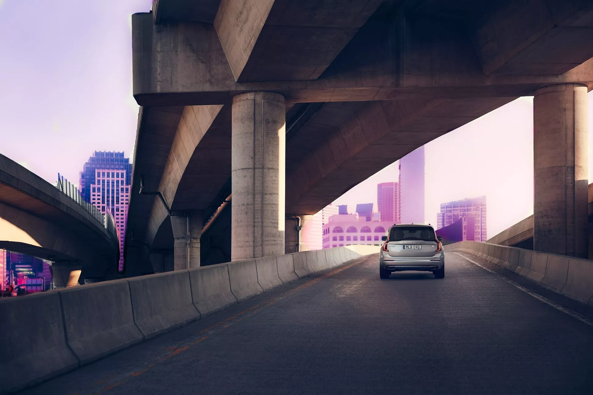 Pohľad zozadu v troch štvrtinách na Volvo XC90 druhej generácie z roku 2015 na zvýšenom diaľničnom moste, proti jemne osvetlenému ružovo-fialovému mestskému panorámu pri svitaní alebo súmraku. Brzdové svetlá auta sú rozsvietené, čo naznačuje spomalenie. SUV má vysokú strechu, zadnú časť s výklopným vekom a aerodynamický dizajn pre efektivitu a štýl, zachytený v rozptýlenom prirodzenom svetle.
