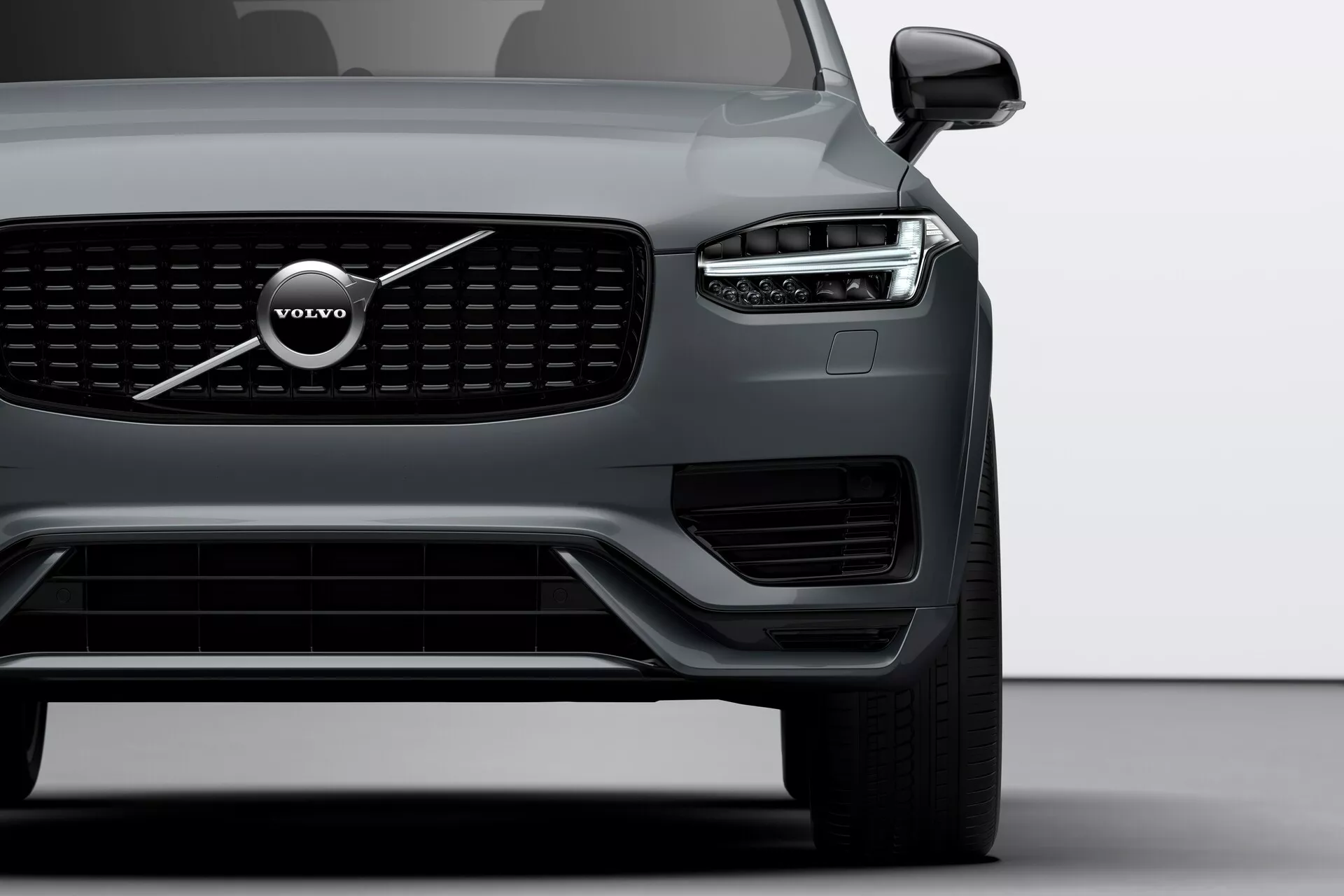 Detailný pohľad na predný uhol Volvo XC90 z roku 2015 (2. generácia) zameraný na ľavú stranu, zvýrazňujúci výraznú mriežku, veľký emblem Volvo a dizajn predných LED svetlometov 'Thorovo kladivo'. Obrázok zahŕňa svetlomet, predné koleso, časť dverí vodiča, viditeľné senzory na spodnom nárazníku a zrkadlo s možnou technológiou sledovania slepého uhla.