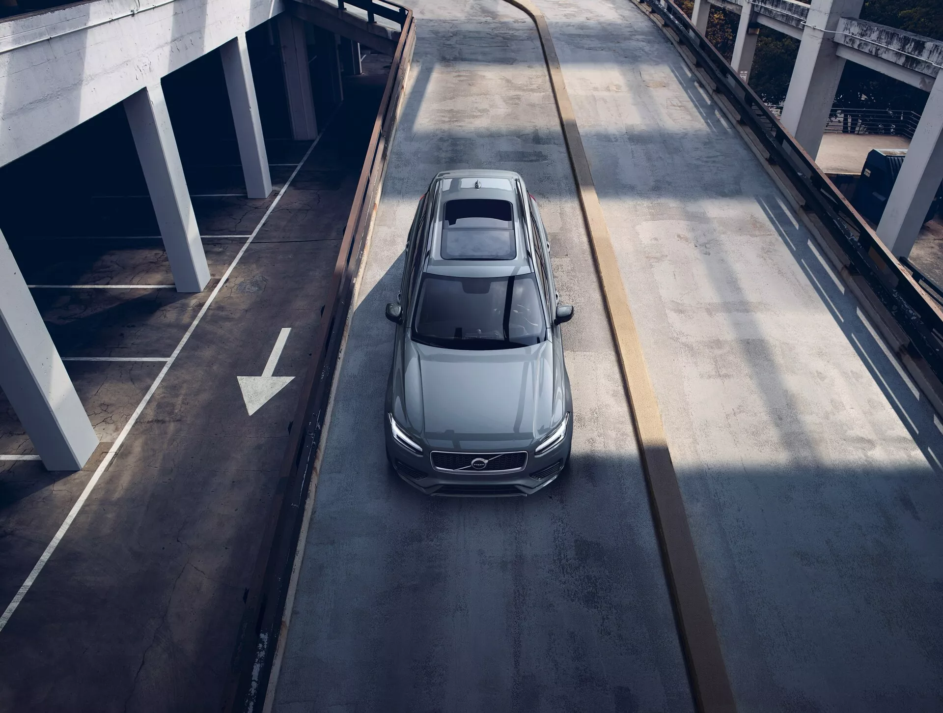 Pohľad z vtáčej perspektívy na sivý alebo strieborný sedan Volvo XC90 z roku 2015, cestujúci po viacúrovňovej mestskej ceste, ukazujúci panoramatickú strechu, aerodynamický dizajn, elegantné LED svetlomety a veľké hliníkové kolesá, čo naznačuje moderné luxusné vozidlo.