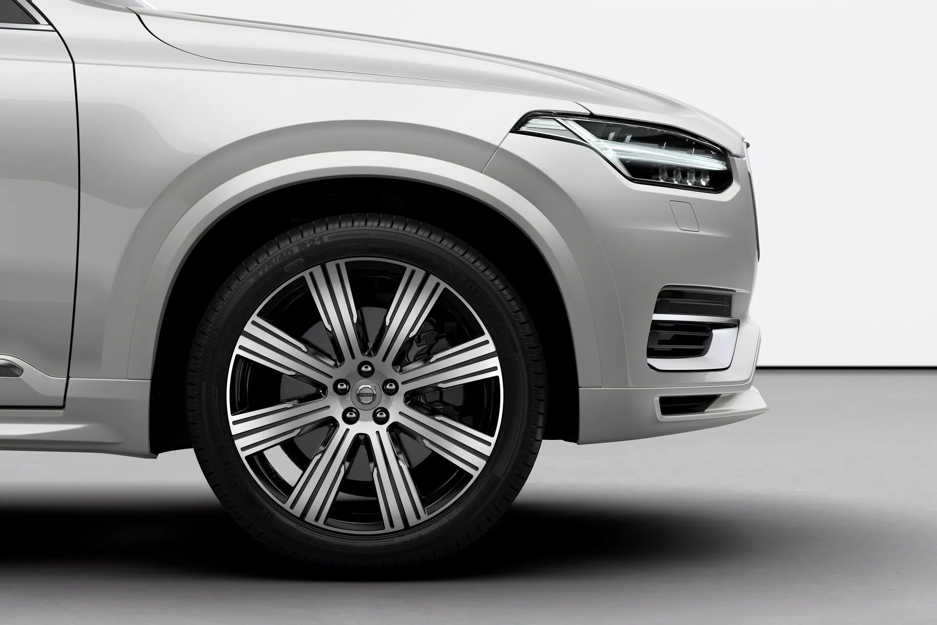 Detailný záber z nízkeho uhlu na predný štvrtinový panel vozidla Volvo XC90 2015 2. generácie, zobrazujúci predné koleso na strane vodiča s viacramenným dizajnom z ľahkej zliatiny, kotúčové brzdy, viditeľný brzdový strmeň a cestnú pneumatiku. Obrázok zdôrazňuje predný nárazník s veľkým prívodom vzduchu, svetlým metalickým lakom a hladkými zakrivenými líniami aerodynamického tvaru.