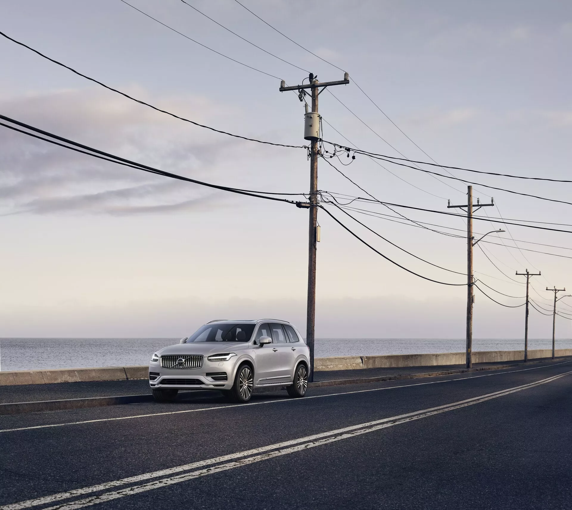 Volvo XC90 druhej generácie z roku 2015, luxusné SUV zaparkované na asfaltovej ceste paralelnej s pobrežím oceánu, zachytené za denného svetla s mäkkým rozptýleným svetlom a dlhými tieňmi. Pohľad z predného uhla zvýrazňuje elegantné dizajnové prvky, vrátane svetlometov, mriežky a kontúr kapoty, svetlosivá alebo bledošampaňská farba, ktorá kontrastuje s tmavou cestou a prírodným pozadím.