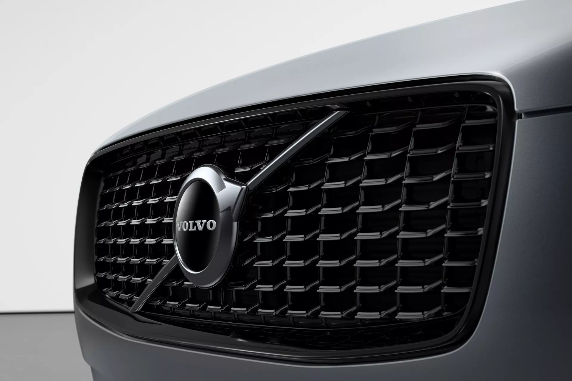 Detailný záber prednej časti vozidla Volvo XC90 z roku 2015 (2. generácia) zameraný na mriežku, ktorá má hustý mriežkový vzor s obdĺžnikovými prvkami a chrómovaný znak Volvo v strede so šípkou smerujúcou šikmo nahor doprava a pohľad na tmavú karosériu vozidla vľavo. Obrázok zdôrazňuje estetiku dizajnu a rozpoznanie značky.