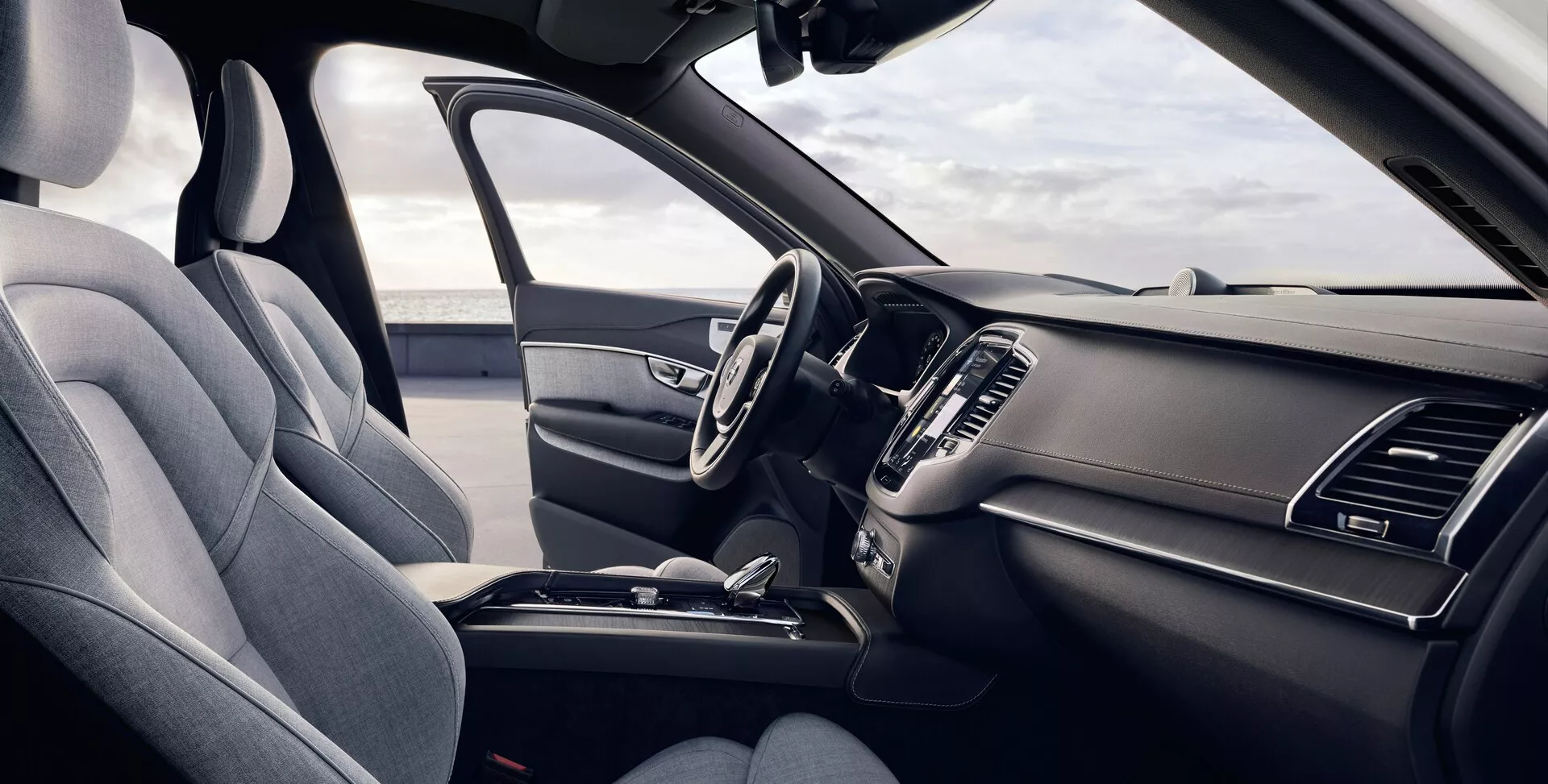 Tento obrázok ukazuje interiér Volvo XC90 druhej generácie z roku 2015 z pohľadu vodiča. Mierne zvýšený uhol ponúka detailný pohľad na predné sedadlá a prístrojovú dosku, na ktorej je lavé sedadlo vodiča čalúnené textúrovanou látkou s viditeľnou opierkou hlavy a bočnými podpormi. Multifunkčný volant má niekoľko ovládacích tlačidiel a elegantná prístrojová doska obsahuje symetrické vzduchové vetráky a čiastočne viditeľný centrálny infotainmentový displej. Stredová konzola ukazuje radiacu páku automatickej prevodovky a ďalšie ovládacie prvky, ktoré zdôrazňujú ergonomický a praktický dizajn.