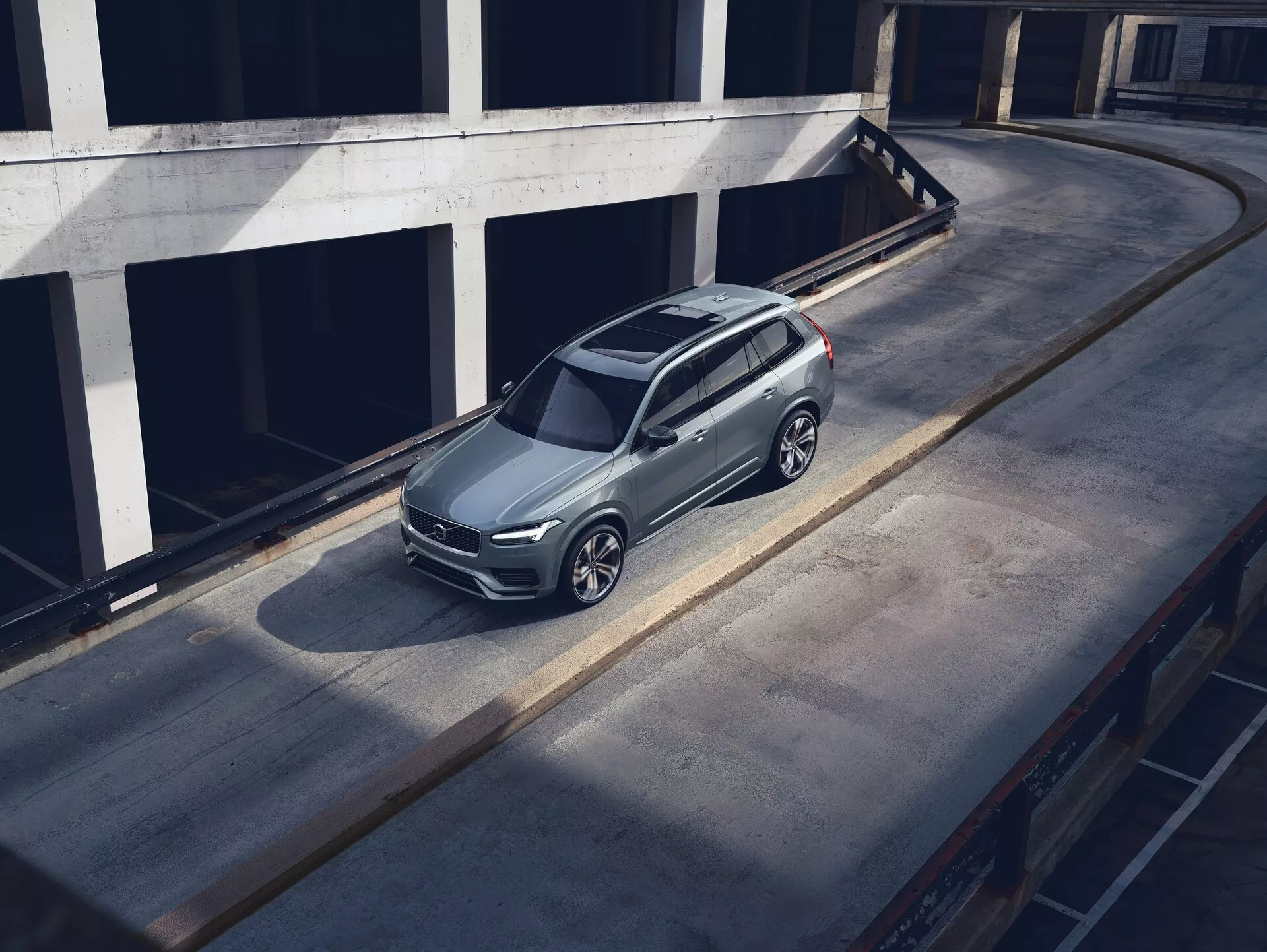 Vysoký pohľad zhora na zaparkované SUV Volvo XC90 z roku 2015 na betónovej ploche, ukazujúce jeho elegantný, moderný dizajn s metalickým lakom, charakteristickou prednou mriežkou, viditeľnými svetlometmi a zložitými kolesami z ľahkých zliatin. Záber zdôrazňuje strechu, kapotu a ľavú stranu vozidla, tieň naznačuje ranné alebo neskoré popoludňajšie svetlo. Prostredie vyzerá ako horné poschodie viacposchodovej parkovacej štruktúry, čo zvyšuje význam vozidla v čistom, mestskom prostredí.