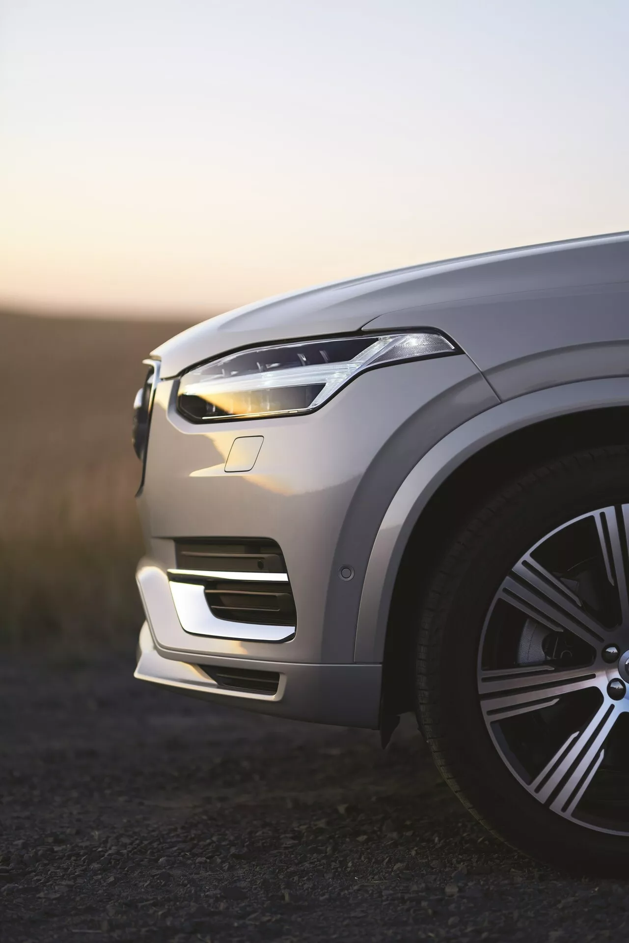 Pohľad na ľavý predný roh luxusného SUV Volvo XC90 2. generácie z roku 2015 za úsvitu, osvetlené LED denné svetlá, kovový lak odrážajúci okolití svetlo, štýlové veľké zliatinové koleso, zložité kontúry kapoty, elegantný dizajn LED svetlometu, integrovaný predný nárazník s nasávačmi vzduchu, viditeľné potenciálne senzory asistenta riadenia, plytká hĺbka ostrosti s rozmazaným pozadím.