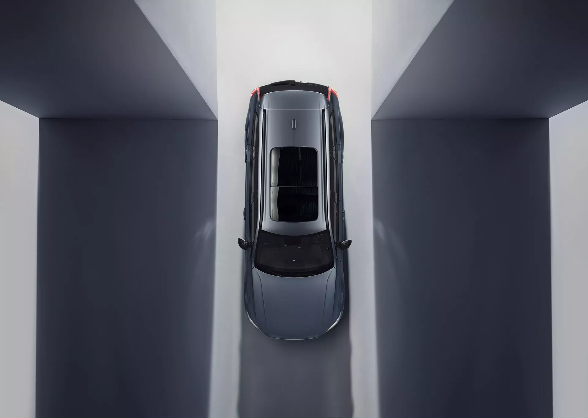 Letecký pohľad na Volvo XC90 2015 (2. generácia) zobrazujúci symetrickú kompozíciu a odrazy prostredia. Centrálna poloha v neutrálnom tónovanom vnútornom prostredí zvýrazňuje strechu vozidla so strešným oknom alebo panoramatickou sklenenou strechou. Viditeľné časti zahŕňajú kapotu, strechu, kufor a hornú polovicu dverí, zdôrazňujúc elegantné línie a krivky naznačujúce luxusný dizajn zameraný na aerodynamiku a priestranné osvetlenie interiéru.