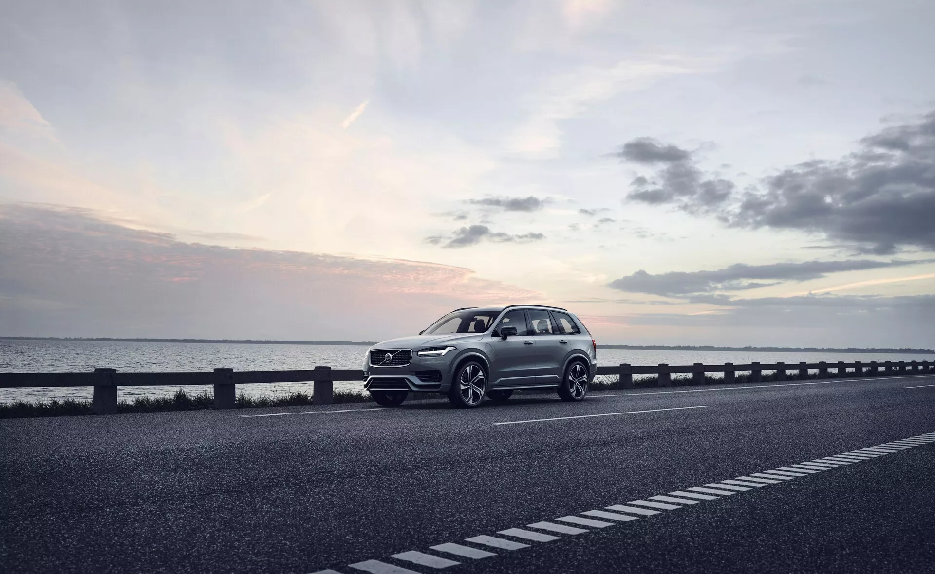 Moderné SUV, konkrétne Volvo XC90 z roku 2015 (2. generácia), zaparkované na pravej strane dvojprúdovej cesty. Vozidlo je zachytené z trojštvrťového predného pohľadu, zobrazujúceho prednú mriežku, svetlomety, kapotu a blatník na strane vodiča. Uhol tiež odhaľuje časť profilu auta, vrátane kolies, dverí vodiča a časti zadku. Kolesá sú mierne otočené doprava, s potenciálne športovými prvkami dizajnu, ako je výrazný nárazník a väčšie nasávacie otvory, čo naznačuje dynamický balík. Fotografia, pravdepodobne urobená za súmraku s mäkkým, rozptýleným svetlom pod mierne zatiahnutou oblohou, zvyšuje pokojnú atmosféru. Dobre definovaná karoséria s viditeľnými líniami charakteru naznačuje luxusný model. Technologické vlastnosti vyplývajúce z obrázka zahŕňajú LED svetlá, pokročilé dizajnérske prvky kolies, sofistikovanú prednú mriežku s možnými senzormi alebo kamerami a aerodynamické prvky. Tento kvalitný obrázok by mohol byť vhodný pre reklamu, zdôrazňujúc auto ako luxusnú voľbu životného štýlu.