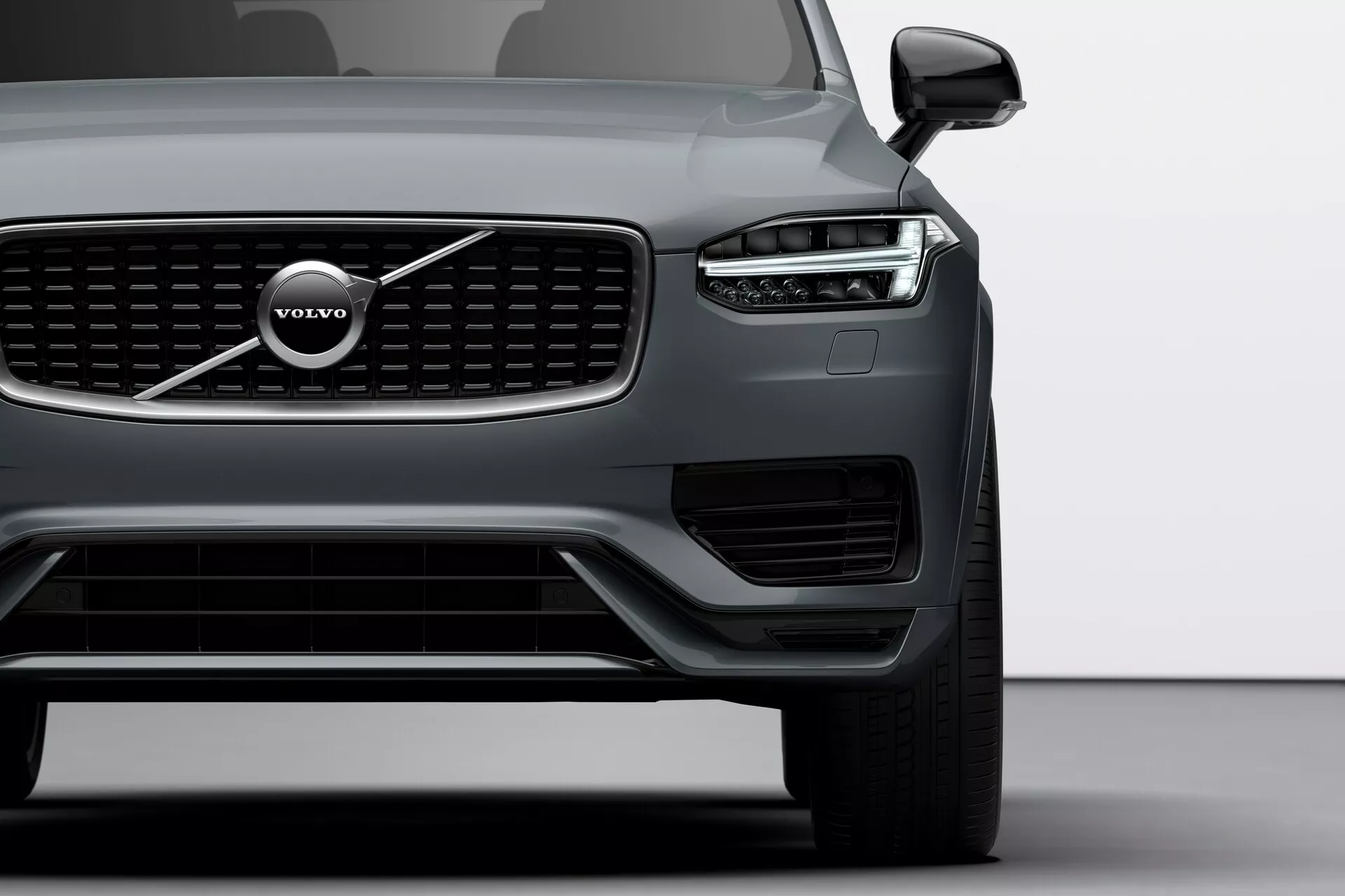 Pohľad na ľavú prednú štvrtinu vozidla Volvo XC90 2015 (druhá generácia), snímka z nízkeho uhla ukazuje ostré, uhlové predné svetlá s charakteristickými dennými LED svetlami v tvare Thorovho kladiva, výraznú prednú mriežku s vertikálnymi lištami a emblémom Volvo, široký spodný prístup vzduchu s dodatočným osvetlením/senzormi, aerodynamické obrysy kapoty, elegantné vonkajšie zrkadlo s integrovaným signalizačným svetlom, čiastočne viditeľné veľké viaclúčové zliatinové koleso a matný povrch karosérie, zdôrazňujúci moderný dizajn a pokročilú technológiu vozidla.