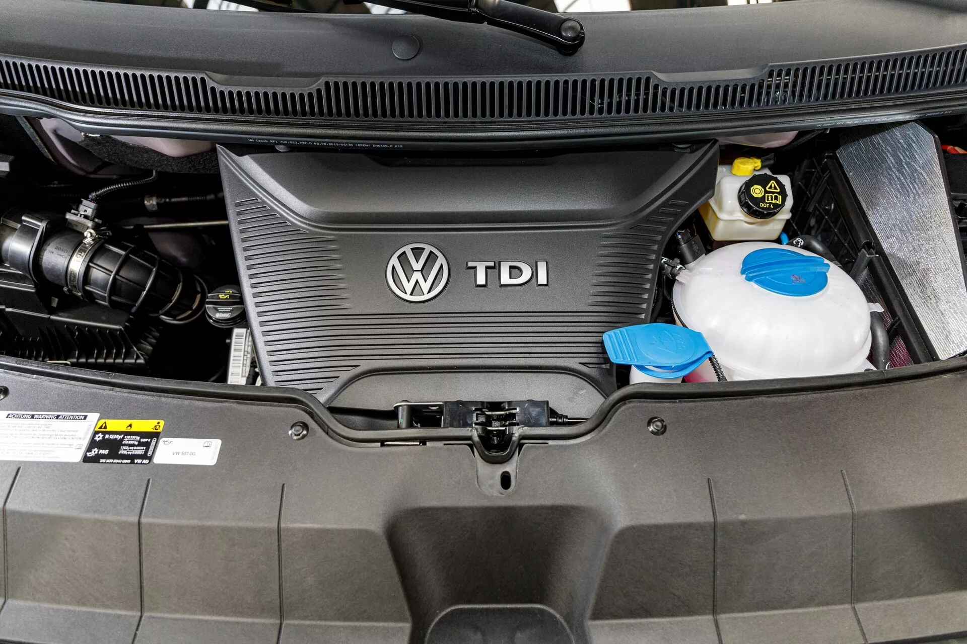 Detailný pohľad na motorový priestor dieselového motora VW Nutzfahrzeuge Transporter 2019 (T6.1 California) s krytom motora TDI s logom Volkswagen, zásobníkom chladiacej kvapaliny a autobatériou