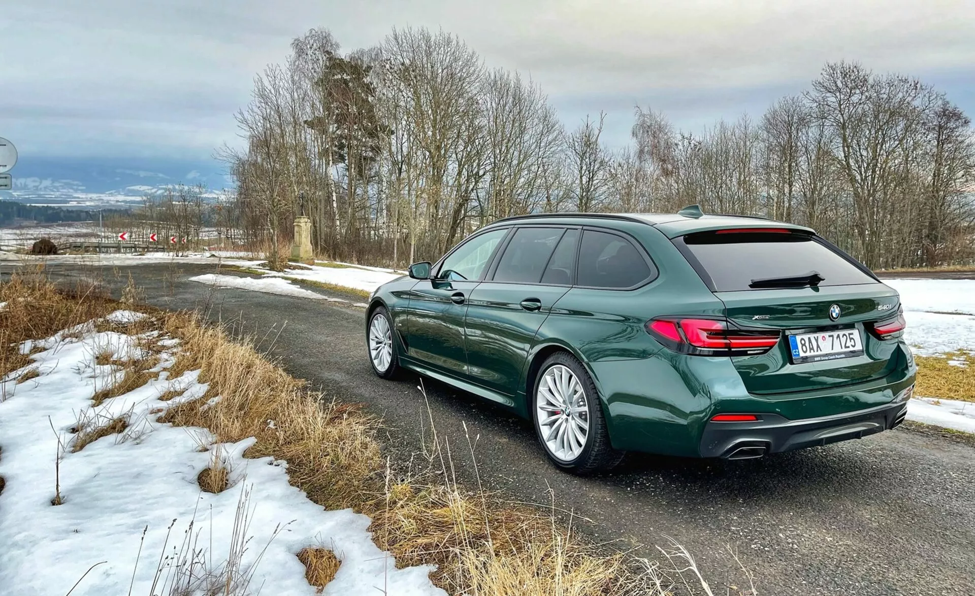 Zadný trojštvrťový pohľad na kombi BMW, ktorý ukazuje zadnú časť vozidla so svetlami v tvare L, ľavý bočný profil vrátane dverí a zakrivenia strechy s nosnými koľajnicami, zliatinové disky s cestnými pneumatikami, v tmavozelenej metalíze, zaparkované na vidieckej ceste s ľahkým snehom, zdôrazňujúce luxus a moderné dizajnové prvky.