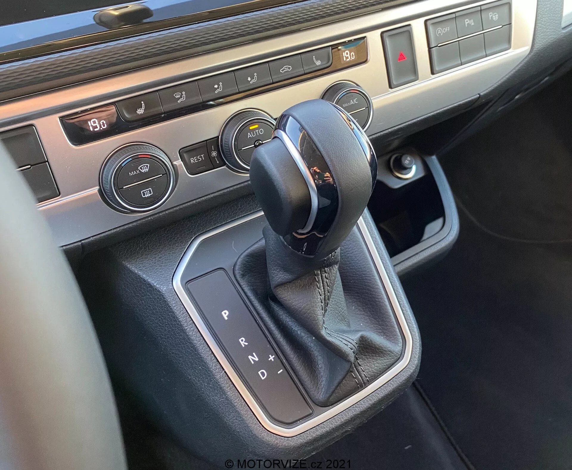 Detailný pohľad na stredovú konzolu vozidla VW Nutzfahrzeuge T6-1 Multivan 2019 zameraný na oblasť radiacej páky. Obrázok zobrazuje automatický volič prevodovky s koženým prešívaním, obklopený kovovým lemovaním, tlačidlá ovládania klimatizácie s displejom teploty a ovládaním vyhrievania sedadiel, ktoré ukazujú moderné vnútorné technológie a prémiové materiály vozidla.
