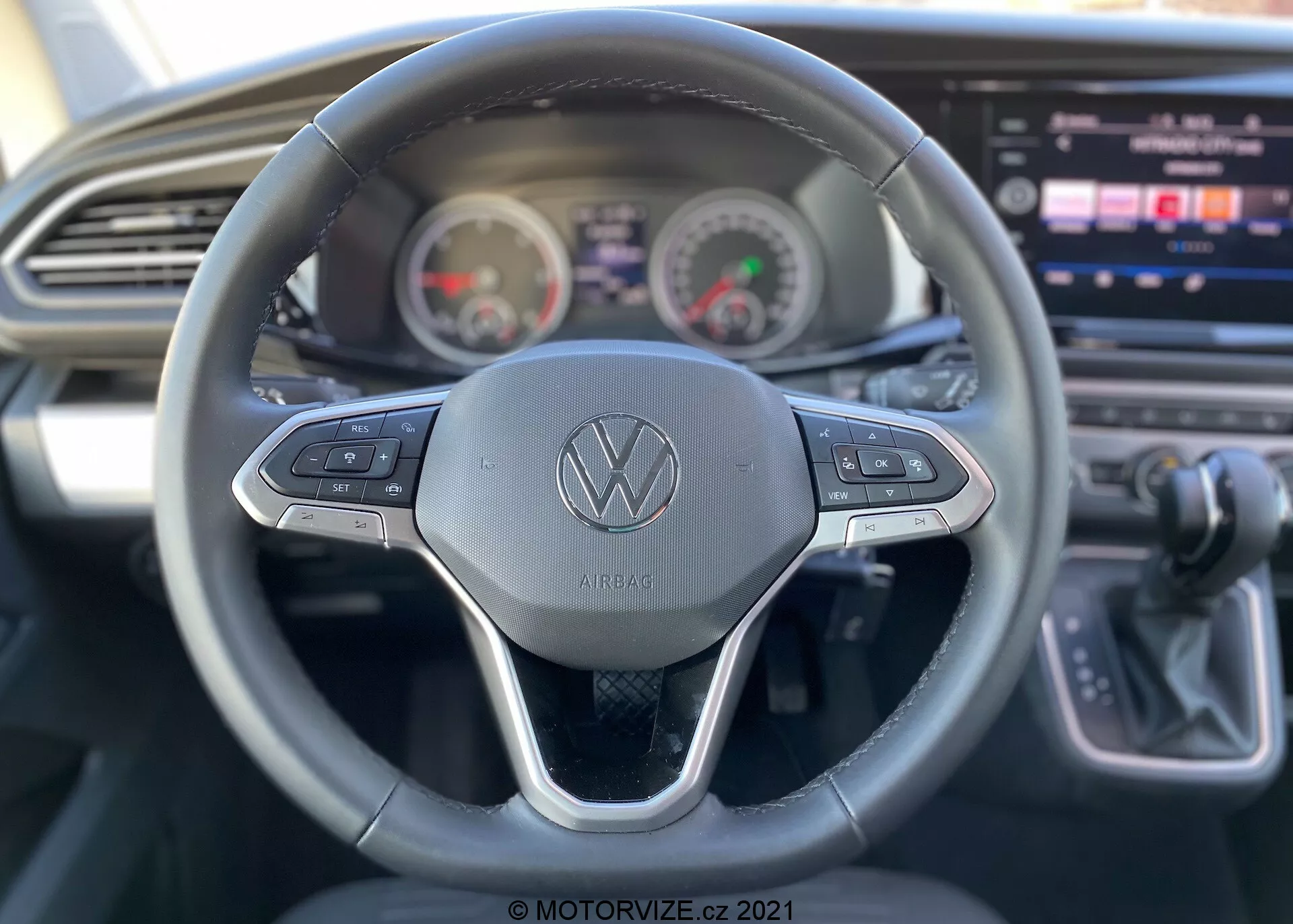 Pohľad na interiér automobilu VW Nutzfahrzeuge Transporter 2019 (T6-1 Multivan) z perspektívy vodiča. Detailný pohľad na volant so značkou Volkswagen a ovládacími tlačidlami pre rôzne funkcie, ako je tempomat a navigácia multimediálnych systémov. Za volantom je panel prístrojov s analógovým tachometrom a otáčkomerom a digitálnym displejom medzi nimi. Čiastočne viditeľná stredová konzola s veľkým dotykovým displejom infotainmentu, spolu s elegantným dizajnom palubnej dosky s vodorovným ventilačným otvorom. Páka prevodovky na pravej strane naznačuje automatickú prevodovku. Obrázok zdôrazňuje použiteľné funkcie so zameraním najmä na volant.