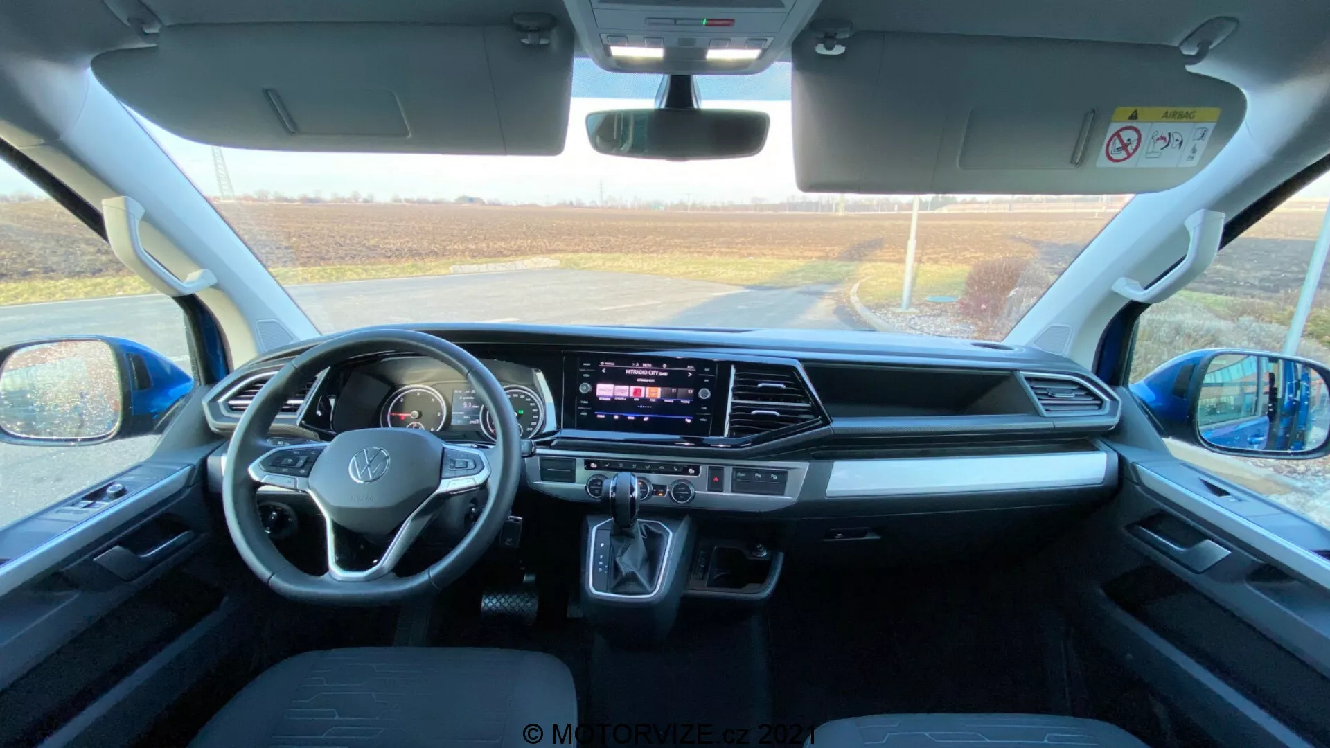 Interiér vozidla VW Nutzfahrzeuge Transporter 2019 (T6.1 Multivan), zachytený z priestoru zadných sedadiel smerom k prednej palubnej doske. Fotografia bola urobená počas dňa, prirodzené svetlo vniká čelným sklom, poskytuje podrobný pohľad na priestor kabíny, predné sedadlá, volant s namontovanými ovládacími prvkami, centrálny dotykový displej infotainmentu, ovládacie tlačidlá a gombíky klimatizácie, automatický radič prevodovky, úložné priestory, konektivné porty, ovládanie dverí a zrkadiel, bezpečnostné prvky a pohodlný dizajn sedadiel.
