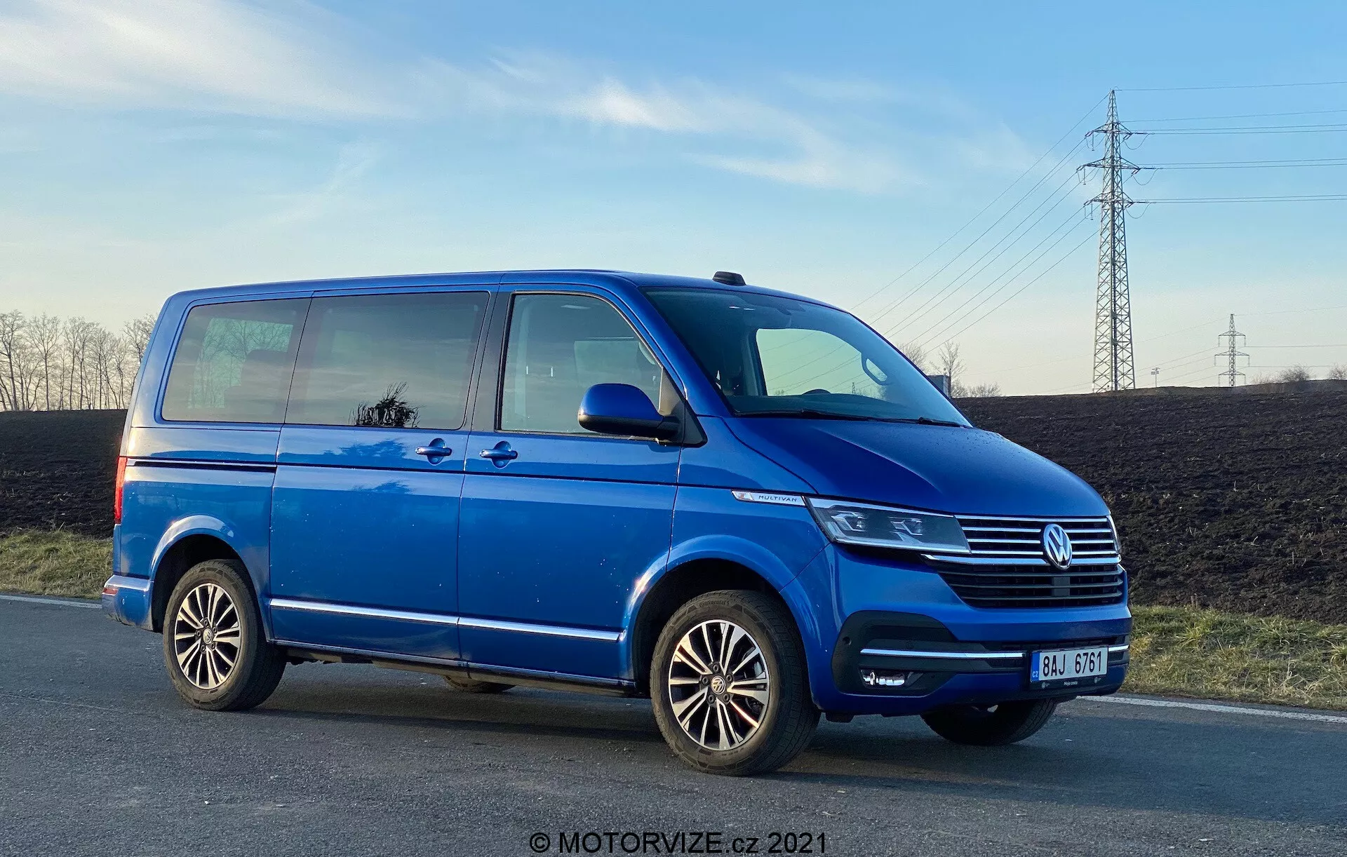 Modrá dodávka Volkswagen Transporter T6, ktorá ukazuje svoj bočný profil z nízkeho uhla, zdôrazňujúc svoju dĺžku a výšku. Obrázok zahŕňa detaily ako posuvné dvere pre pasažierov, predné dvere pre pasažierov, zadné koleso na strane pasažiera, viacramenné zliatinové disky a čiastočné pohľady na svetlomet, mriežku chladiča Volkswagen s emblémom a predný nárazník. Fotografia bola zhotovená v neskorom popoludní a ukazuje svetlú výšku, pričom odráža nebo a okolie v oknách, s čistou oblohou a elektrickými vedeniami v pozadí.