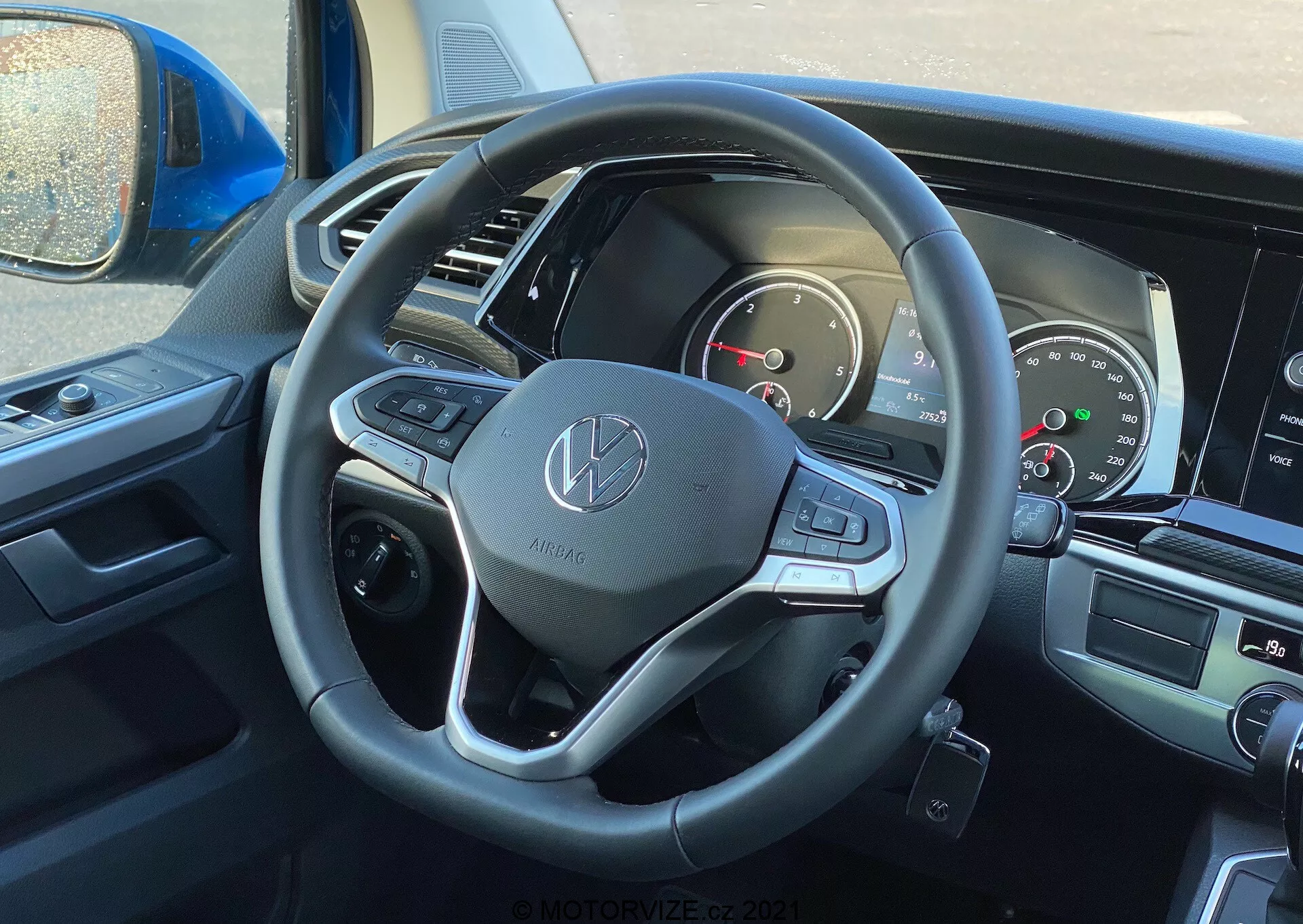 Pohľad do interiéru vozidla VW Nutzfahrzeuge Transporter 2019 (T6-1 Multivan) z perspektívy vodiča so zameraním na volant a prístrojovú dosku. Volant má emblém Volkswagen a je vybavený koženým poťahom, multifunkčnými tlačidlami a radiacimi pákami. Čiastočne viditeľný prístrojový panel zahŕňa analógové ciferníky a digitálny displej. Centrálna infotainment obrazovka a ovládanie klimatizácie sú slabo viditeľné, čo naznačuje moderné multimediálne a komfortné funkcie.