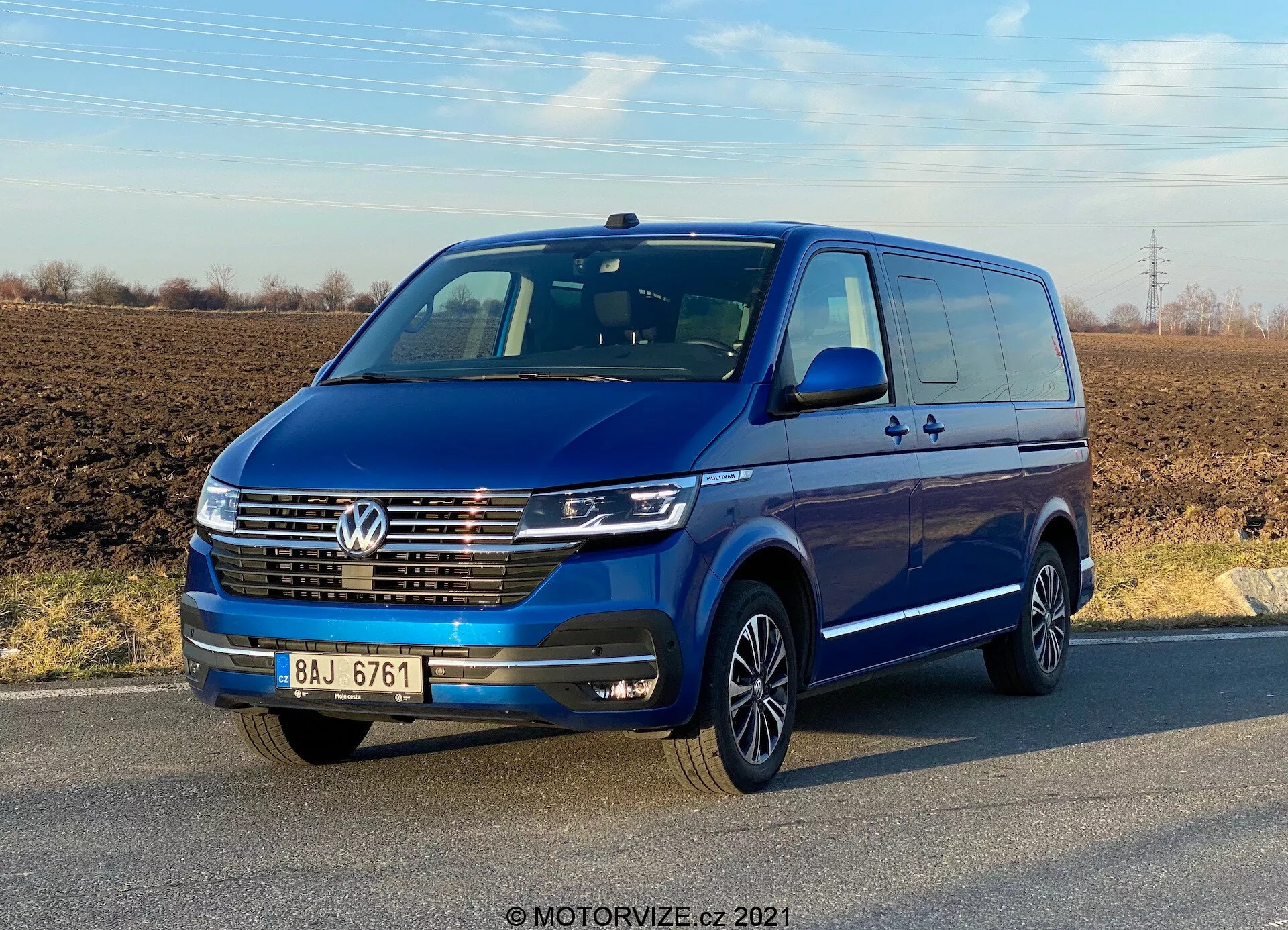 Čelný pohľad na modrý Volkswagen Transporter T6.1 Multivan 2019 pod jasnou oblohou. Vozidlo má znak Volkswagen na maske, LED denné svietenie, chrómovanú lištu na prednom grile a viac ramenné zliatinové kolesá. Zrkadlá a nárazníky vo farbe karosérie naznačujú vyššiu špecifikáciu modelu. Sú viditeľné dvere a kľuky vrátane zadných posuvných dverí, čo naznačuje kapacitu pre viac cestujúcich alebo náklad.