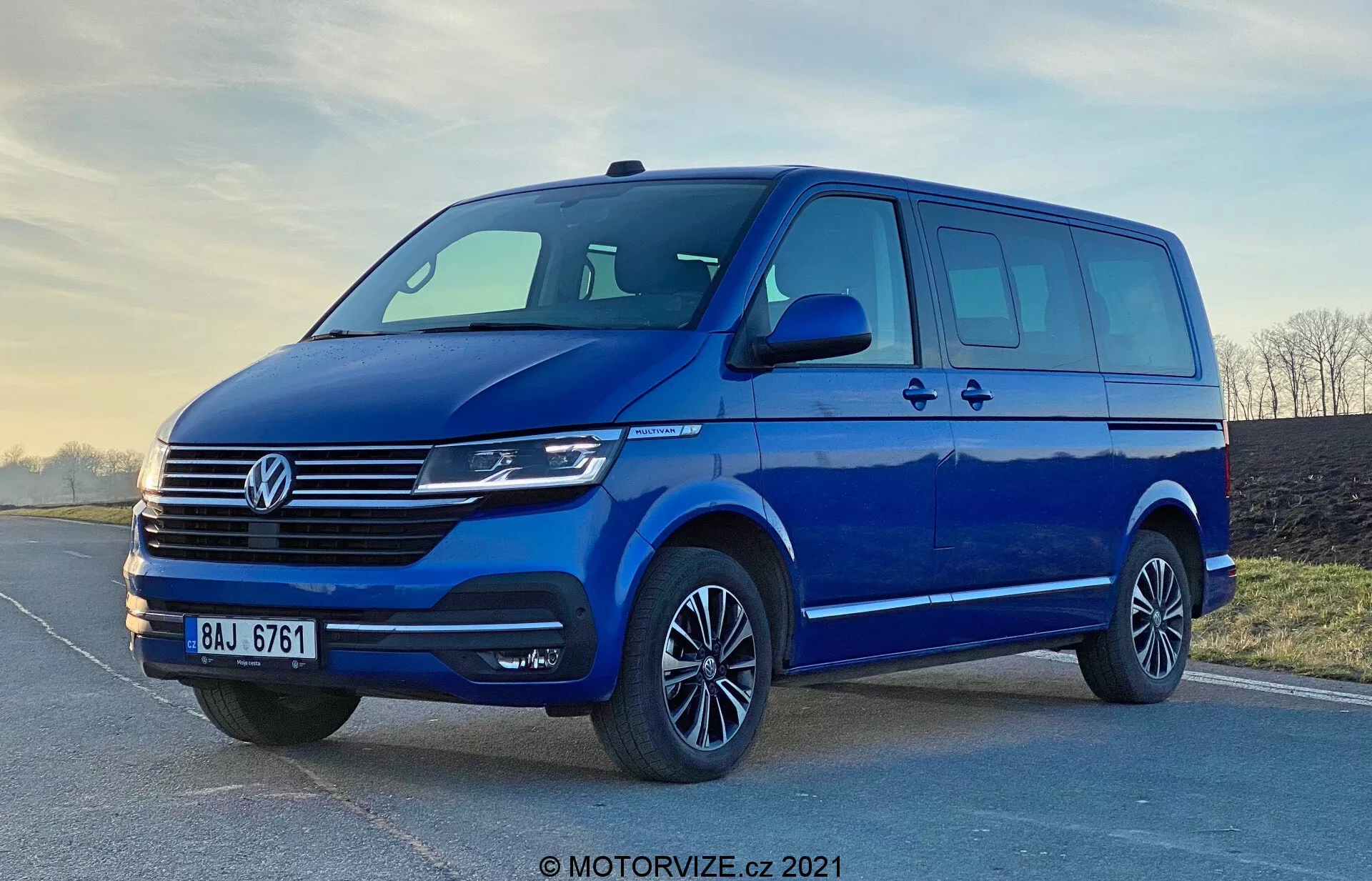 Obrázok modrého Volkswagen Transporter 2019 T6.1 Multivan, odfotografovaný z nízkeho uhla, aby vynikol jeho vzrast a veľkosť. Predný trojštvrťový pohľad ukazuje predný a bočný dizajn, vrátane charakteristickej mriežky Volkswagen s horizontálnymi líniami, centrálneho loga, moderných svetlometov pravdepodobne s LED alebo Xenon technológiou a elegantného predného nárazníka so širokým sáním vzduchu. Aerodynamické bočné zrkadlá s integrovanými smerovkami, dostatok okenného priestoru s posuvnými bočnými dverami, zliatinové kolesá s nízkoprofilovými pneumatikami a mäkké osvetlenie zdôrazňujú praktickosť a estetický vzhľad dodávky. Vodotlač 'MOTORVIZE.cz 2021' naznačuje, že obrázok pochádza z roku 2021.