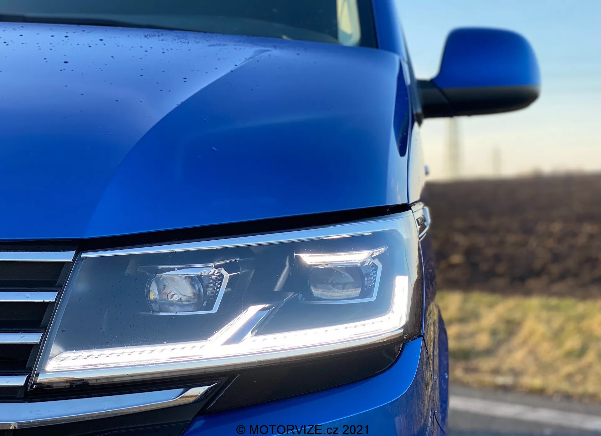 Detailný záber z nízkeho uhla na prednú ľavú časť modrého Volkswagenu Nutzfahrzeuge Transporter 2019 (T6.1 Multivan), ktorý zdôrazňuje ostré a uhlové moderné LED svetlomety s denným osvetlením, časť chrómovanej mriežky, ako aj obrys kapoty a spätné zrkadlo. Fotografia s nízkou hĺbkou poľa zdôrazňuje svetlomet a okolie karosérie, vyzdvihuje pokročilú svetelnú technológiu, súčasný dizajn a kvalitu laku, doplnené o kvapky dažďa, ktoré naznačujú podrobnú vonkajšiu úpravu.