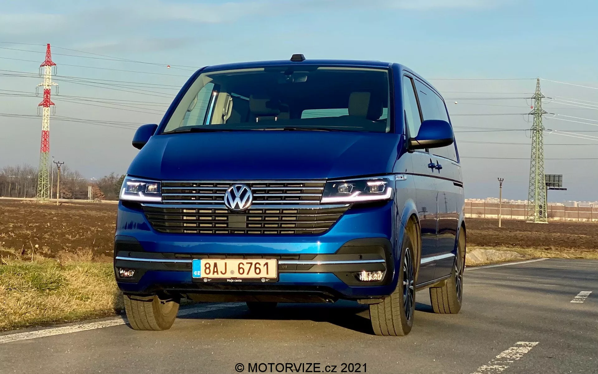 Modrý Volkswagen T6-1 Multivan 2019 zachytený z nízkeho predného uhla zvýrazňujúceho mriežku, LED svetlomety s dennými svetlami, emblém Volkswagen, širokú mriežku, dvojfarebný nárazník a liate kolesá; zaparkovaný pri ceste s jasnou oblohou, vzdialenou zeleňou, elektrickým stĺpom a infraštruktúrou v pozadí.