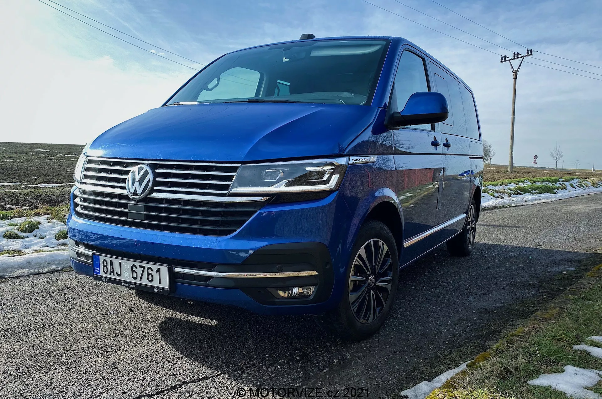 Modrý Volkswagen T6.1 Multivan Transporter 2019 zachytený z nízkeho trojštvrťového predného uhla, zobrazujúci prednú časť vozidla, masku chladiča s výrazným logom Volkswagen, horizontálnymi chrómovými pruhmi, hranatými LED svetlometmi s dennými svetlami, predný nárazník s integrovanými hmlovými svetlami a nasávacími otvormi, dynamickú vykrojenú líniu tiahnucu sa po bokoch, viacramenné hliníkové kolesá, integrované bočné zrkadlá s blinkermi, zatmavené okná. Vozidlo je zaparkované na mokrej ceste vedľa zasneženej trávy, s vidieckym pozadím obsahujúcim stĺpy elektrického vedenia a otvorenú oblohu.