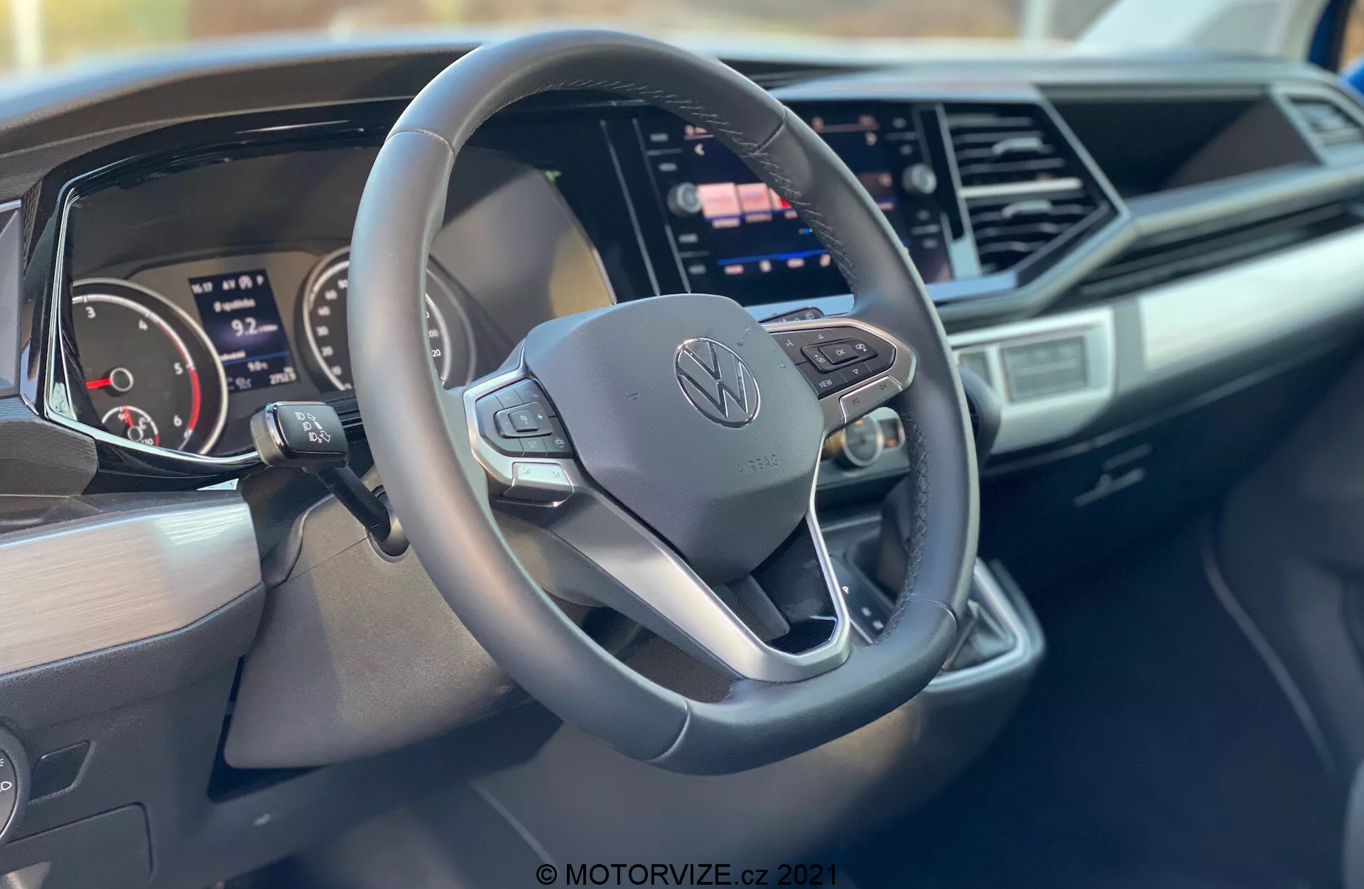 Interiérový záber vozidla Volkswagen Nutzfahrzeuge Transporter 2019 (T6-1 Multivan) so zameraním na volant a prístrojovú dosku. Detailný záber z pohľadu spolujazdca zvýrazňuje trojramenný, dolu sploštený volant s ovládacími tlačidlami a logom Volkswagen v strede, digitálny prístrojový panel, veľký dotykový displej infotainmentu, elegantný dizajn prístrojovej dosky s ostrými akcentmi a branding na strane spolujazdca prístrojovej dosky.