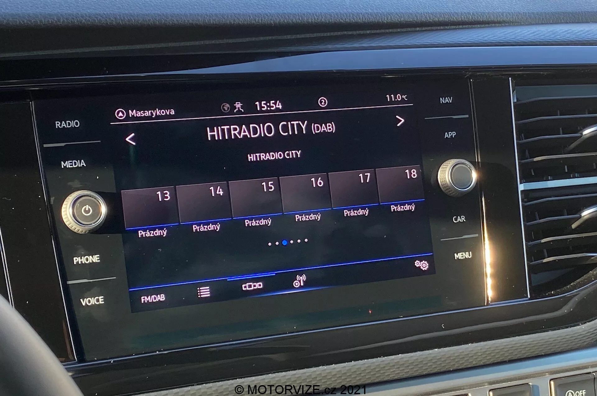 Detailný čelný záber na obrazovku infotainmentového systému vo VW-Nutzfahrzeuge Transporter 2019 (T6-1 Multivan). Obrázok zobrazuje digitálnu rádiostanicu 'HITRADIO CITY' so zobrazenými rôznymi dotykovými možnosťami a funkčnými tlačidlami ako 'PHONE', 'VOICE' a 'MENU'. Displej ukazuje čas '15:54' a vonkajšiu teplotu '11.0°C' s ikonami pre užívateľské profily, nastavenie vozidla a silu signálu. Obrazovka je integrovaná do strednej časti palubnej dosky, obklopená hmatovými tlačidlami a ovládacími gombíkmi.