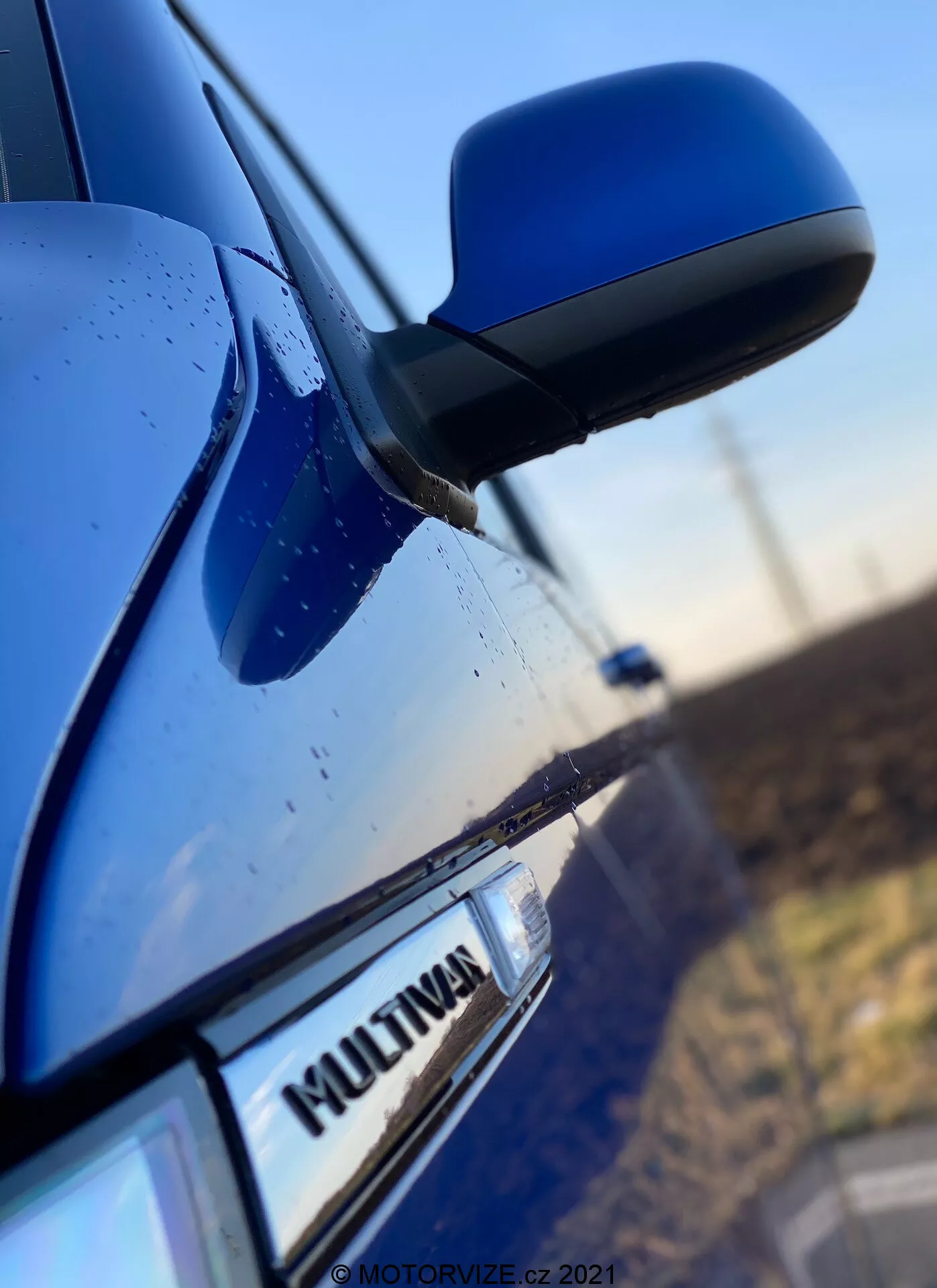 Detailný záber na bočnú časť modrého VW Nutzfahrzeuge Transporter 2019 (T6-1-Multivan), na ktorom je vidieť časť predných dverí spolujazdca, bočné zrkadlo a časť zadných dverí. Pozornosť je zameraná na odznak 'MULTIVAN' na aute, pričom elegantné a aerodynamické bočné zrkadlo je mierne rozmazané. Kryt zrkadla zodpovedá farbe karosérie a na povrchu sú viditeľné kvapky vody, čo naznačuje odolnosť voči poveternostným vplyvom alebo nedávny dážď.