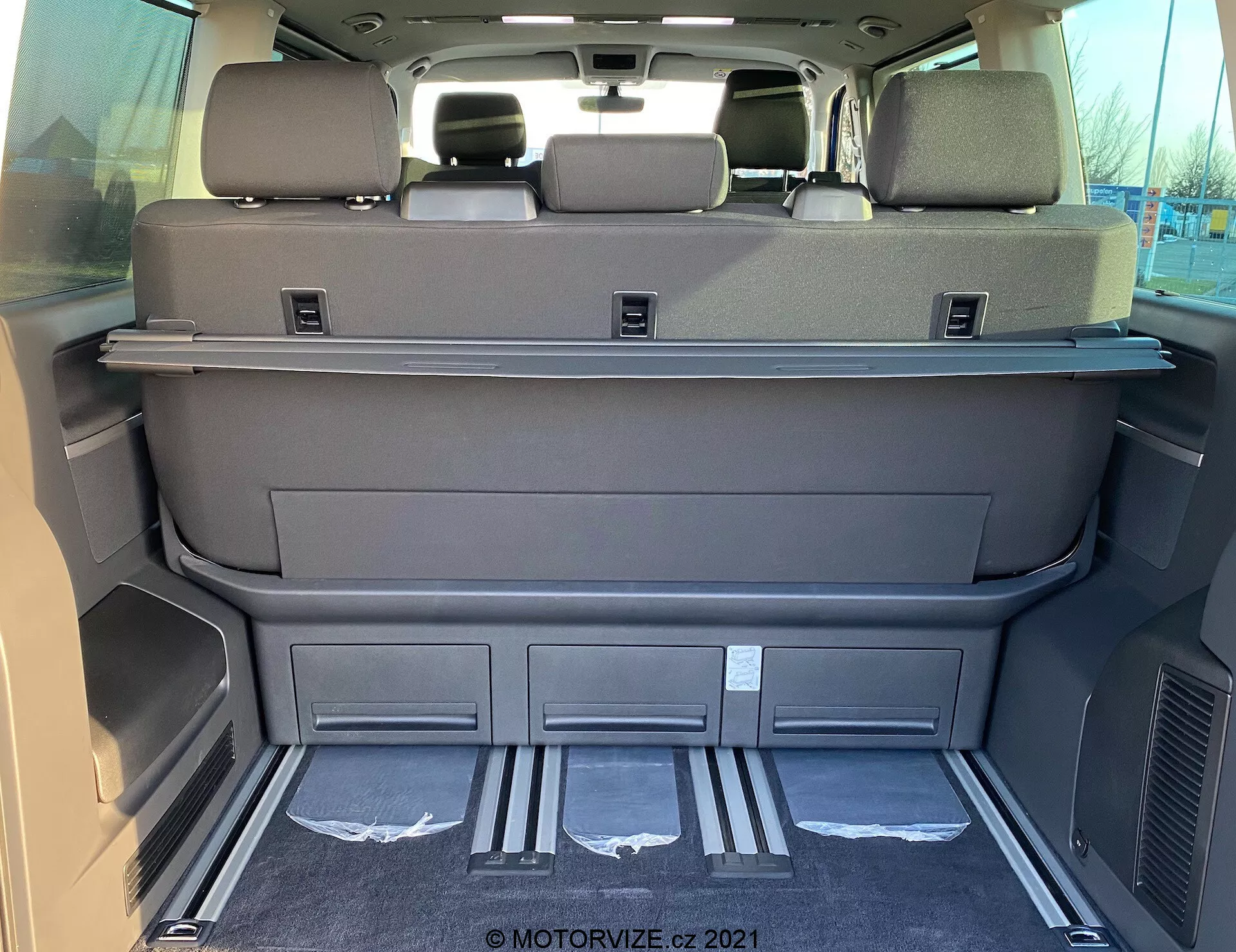 Pohľad na interiér modelu VW Nutzfahrzeuge Transporter T6.1 Multivan z roku 2019 z nákladového priestoru zozadu, ukazujúci priestrannú oblasť so sklopenými sedadlami tretej rady, tmavosivej alebo čiernej farby, policou na krytie nákladu, hlavovými opierkami na zadných sedadlách, kovovými vstupnými krytmi, tvarovanými priehradkami, vzduchovými otvormi, čistým koberecom a možným elektronickým ovládaním sedadiel alebo zadných dverí.