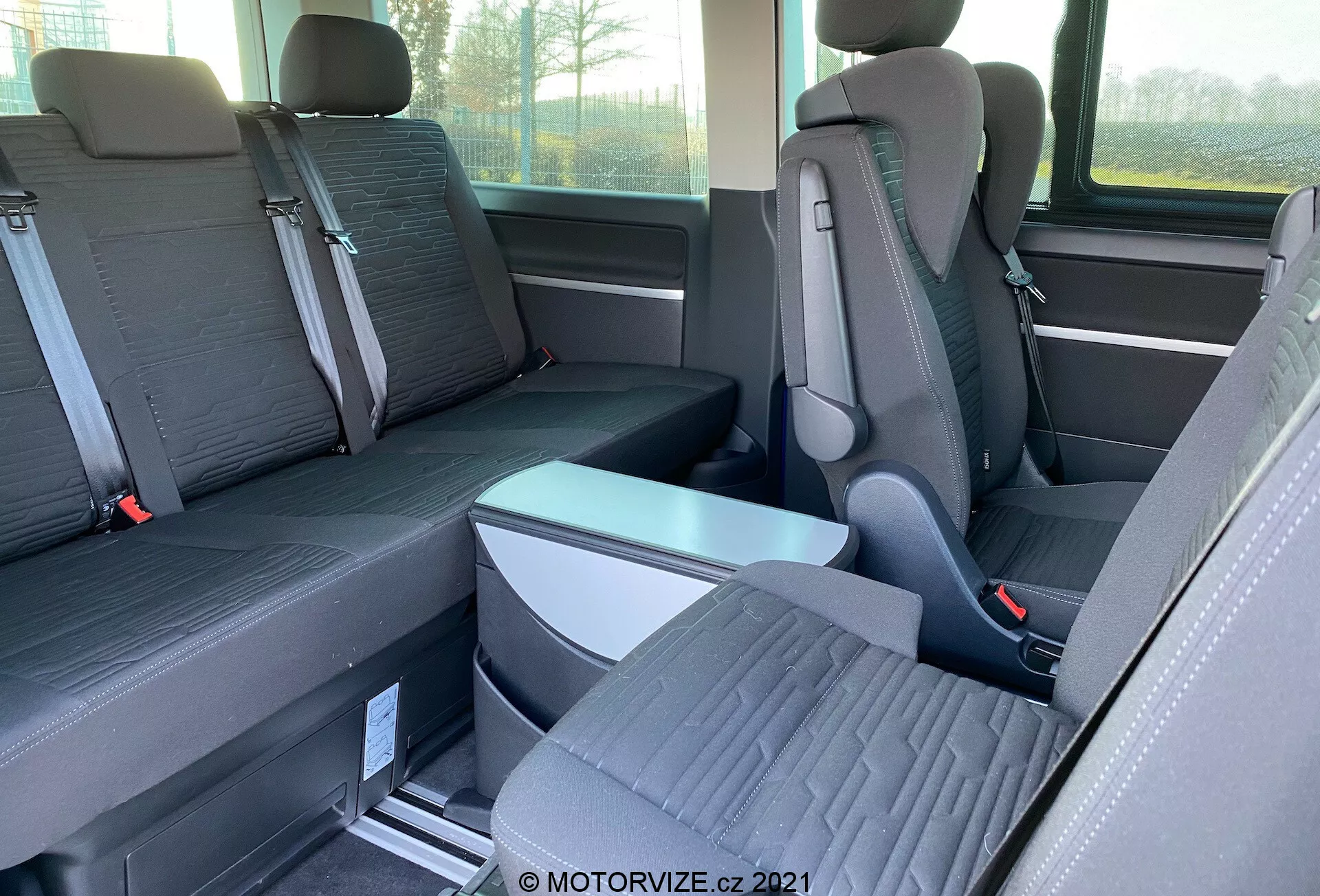 Pohľad do interiéru VW-Nutzfahrzeuge Transporter 2019 (T6.1 Multivan) so zameraním na druhý a tretí rad sedadiel. Fotorgafované z boku vo výške očí, obraz ukazuje jednotlivé sedadlá s výraznými opierkami hlavy v druhom rade a lavicu pre troch cestujúcich v treťom rade. Sedadlá sú z dvojfarebnej látky a podlaha obsahuje kovové koľaje pre prestaviteľné sedadlá. Viditeľná je sklopná opierka a pracky na bezpečnostné pásy. Čiastočne viditeľné je aj okno a bočné úložné priestory.