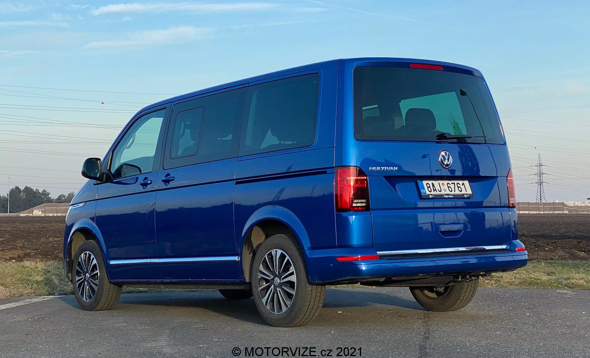 Modrá dodávka VW-Nutzfahrzeuge Transporter 2019 (T6-1 Multivan) z pohľadu z trištvrtečného zadného uhlu. Obrázok zachytáva zadné svetelné klastre, zadné okná a zadné dvere, ako aj bočný profil s dverami pre cestujúcich a oknami. Dodávka zaberá veľkú časť rámu na úrovni očí, odhaľujúc rozdelené zadné dvere a niekoľko bočných okien, čo naznačuje model pre cestujúcich. Zliatinové kolesá, nárazníky vo farbe karosérie a bočné zrkadlá naznačujú vysokú úroveň výbavy. Osvetlenie naznačuje dennú dobu s jasnou oblohou a ostrými tieňmi pod vozidlom.