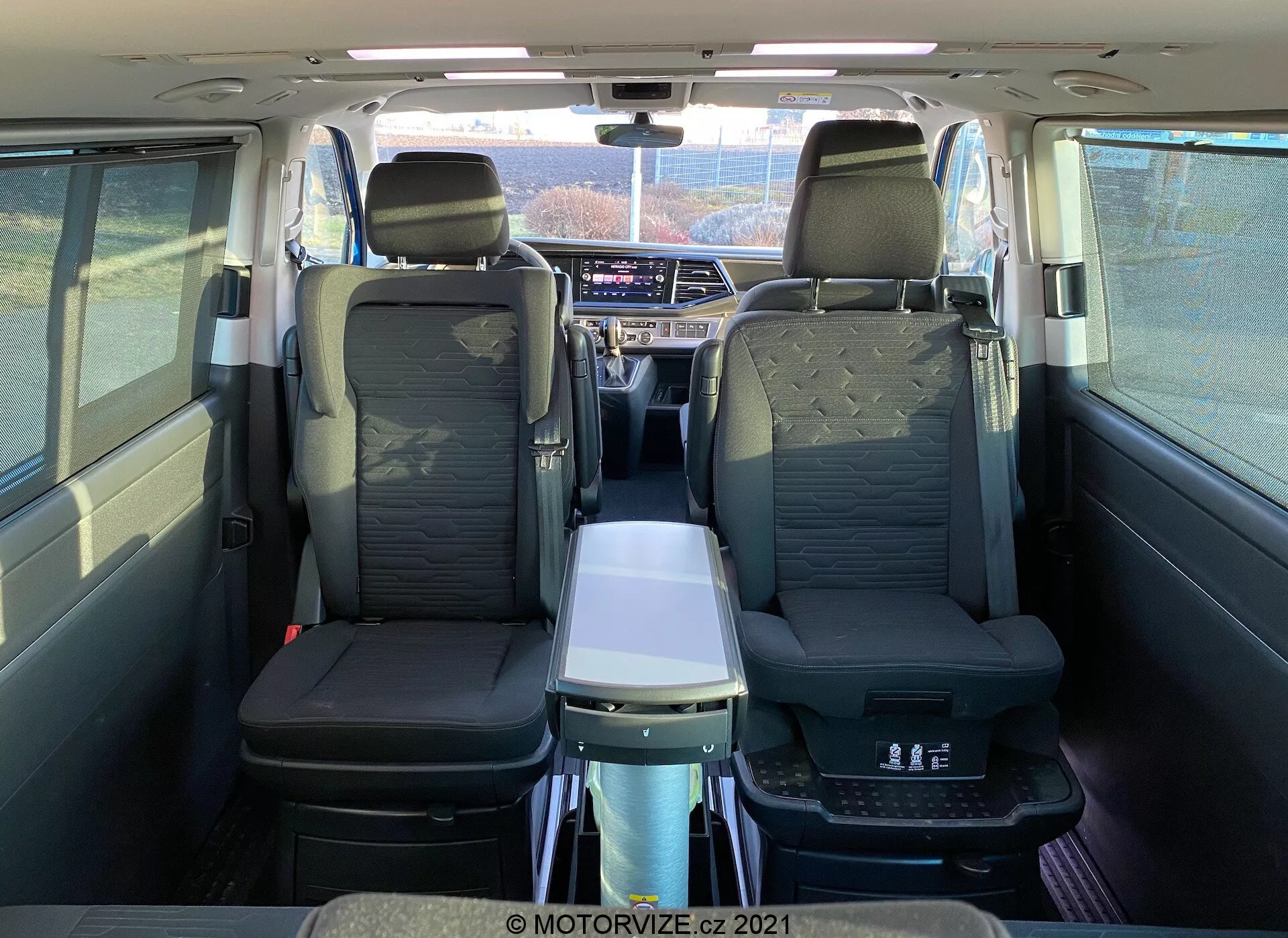 Interiér VW Nutzfahrzeuge Transporter T6-1 Multivan 2019 z pohľadu zozadu, zobrazujúci kožené sedadlá, opierky na ruky v druhej rade, skladací stôl, panoramatickú strechu, individuálne vetracie otvory a pokročilý infotainment systém; zvýrazňuje pohodlie cestujúcich a priestrannosť kabíny.