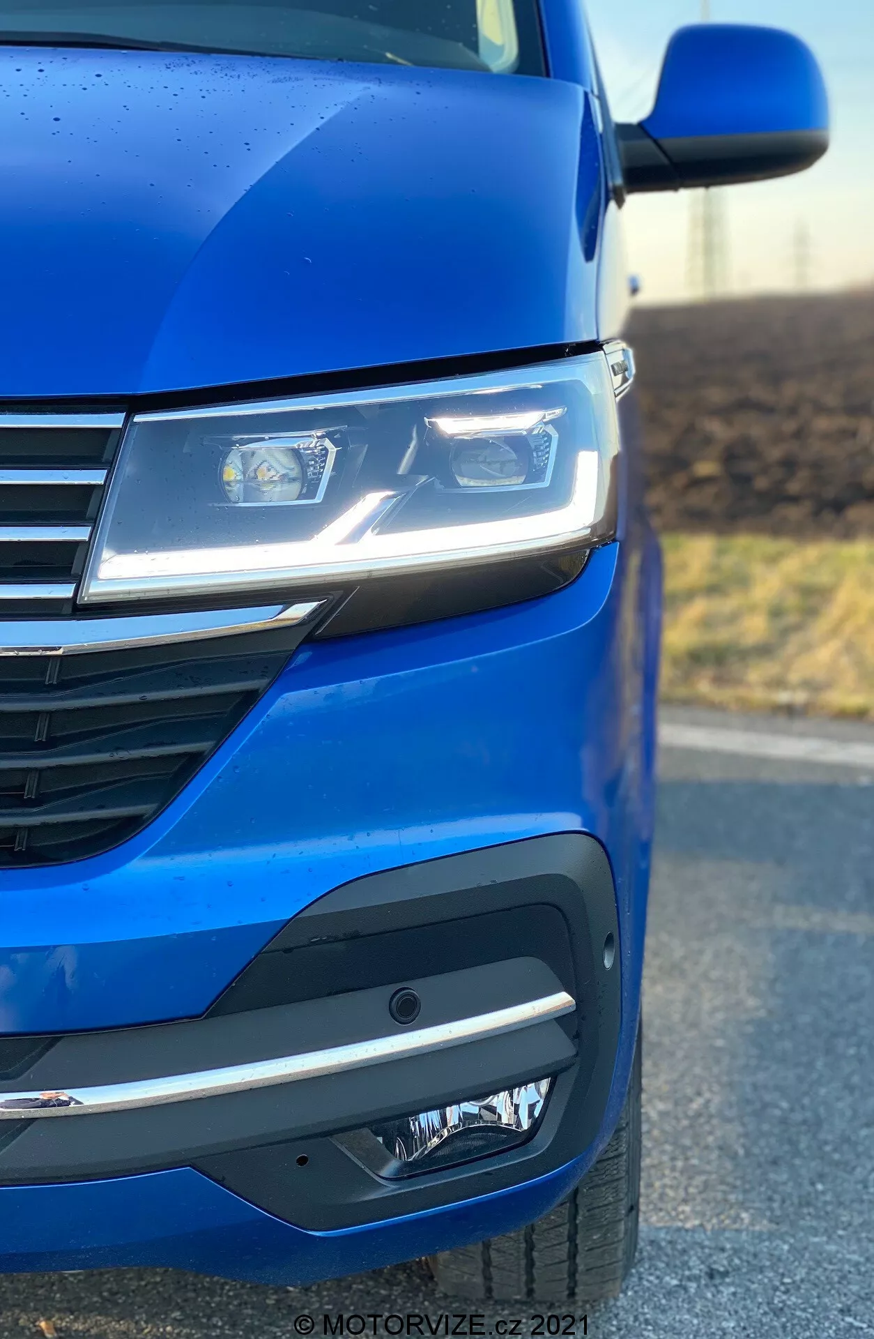 Detailný záber na prednú ľavú stranu VW Nutzfahrzeuge Transporter 2019 (T6.1 Multivan) zdôrazňujúci metalickú modrú farbu, moderné LED svetlomety, moderný dizajn mriežky s horizontálnymi líniami a sofistikovaný predný nárazník s hmlovými svetlami alebo snímačmi. Uhol fotografie zdôrazňuje aerodynamiku a dizajnové prvky auta.