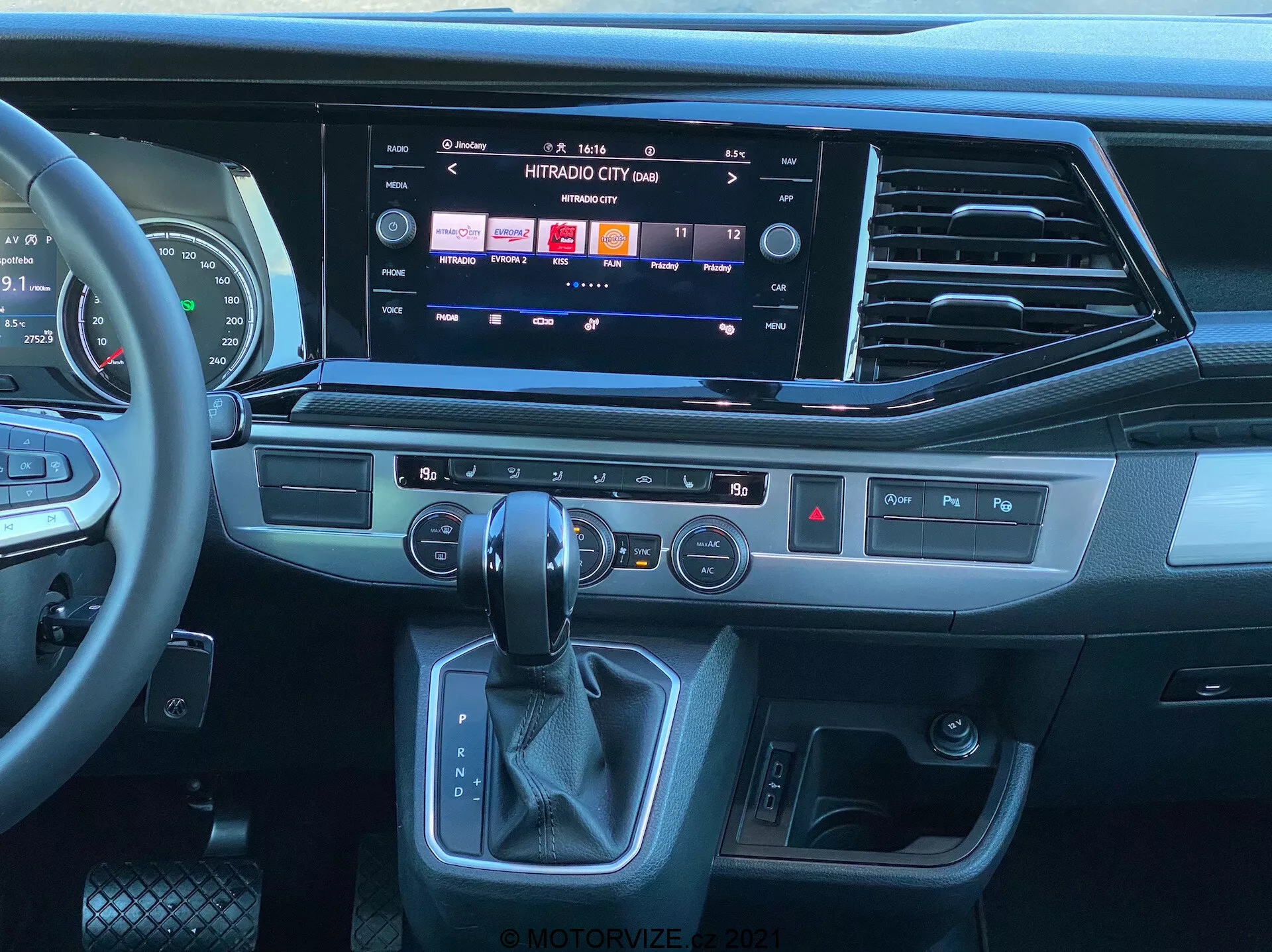 Pohľad na interiér vozidla VW Nutzfahrzeuge Transporter 2019 T6.1 Multivan z perspektívy sedadla spolujazdca, ukazujúc modernú prístrojovú dosku a stredovú konzolu. Multifunkčný kožený volant s ovládaniami médií, veľký dotykový infotainment systém zobrazujúci možnosti médií a konektivity, elegantné ventilačné otvory, hmatové tlačidlá pre rýchle funkcie, panel ovládania klímy s dvojzónovou klímou, ergonomická páka automatickej prevodovky a tlačidlo štart/stop bez kľúča. Dizajn kombinuje tmavé plasty, ozdobné prvky vyzerajúce ako brúsený kov a materiály podobné koži, čo zdôrazňuje moderný a prémiový pocit.