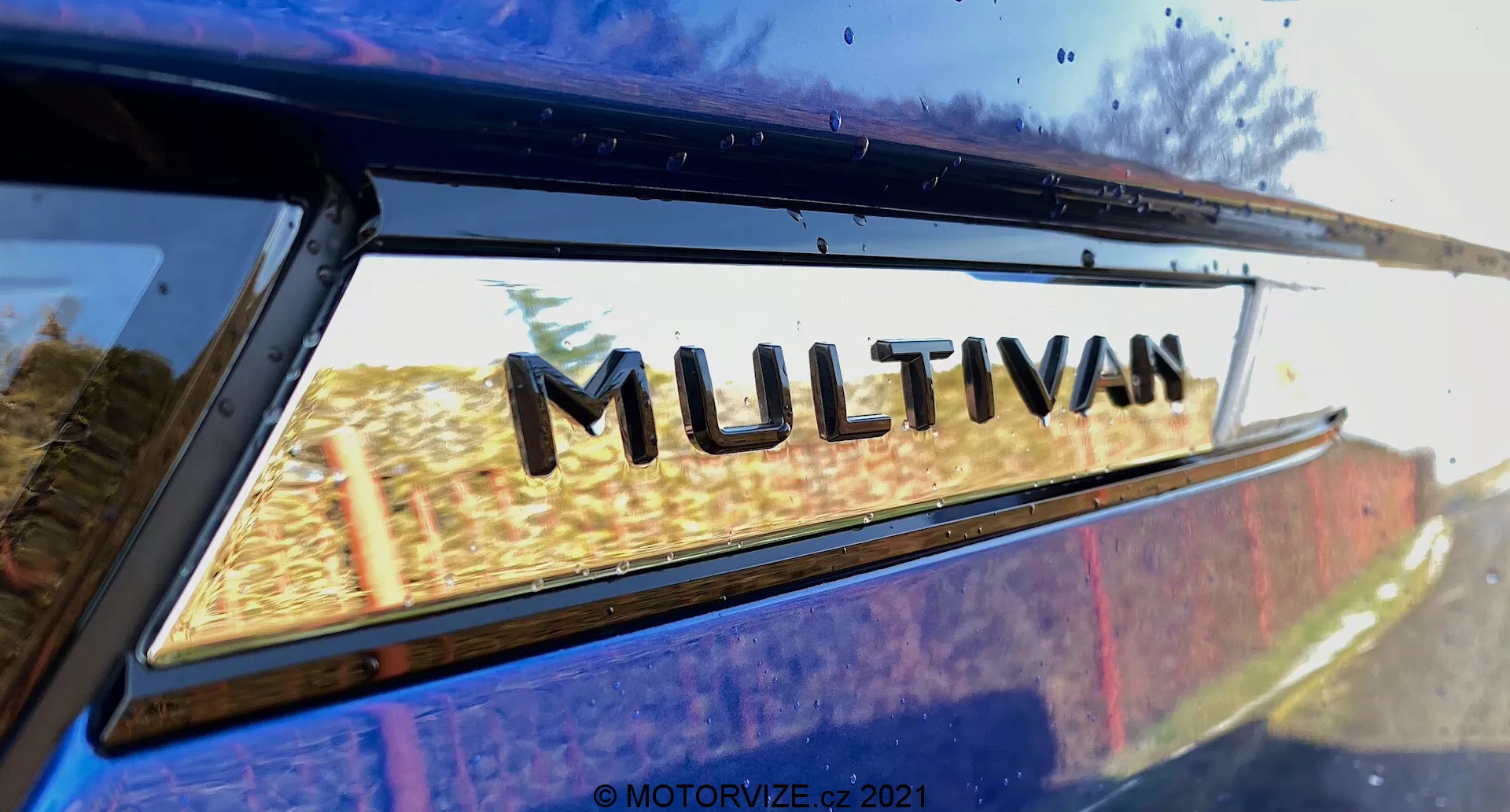 Detailný pohľad na časť VW Nutzfahrzeuge Transporter 2019 (T6-1 Multivan) so zameraním na odznak 'MULTIVAN'. Obrázok je urobený z nízkeho zadného uhlu, mierne nahor, kde sa zadné okno stretáva s okrajom strechy. Jemné odrazy na okne naznačujú vonkajšie prostredie. Tento tesne orezaný obrázok zdôrazňuje označenie modelu výrobcu.