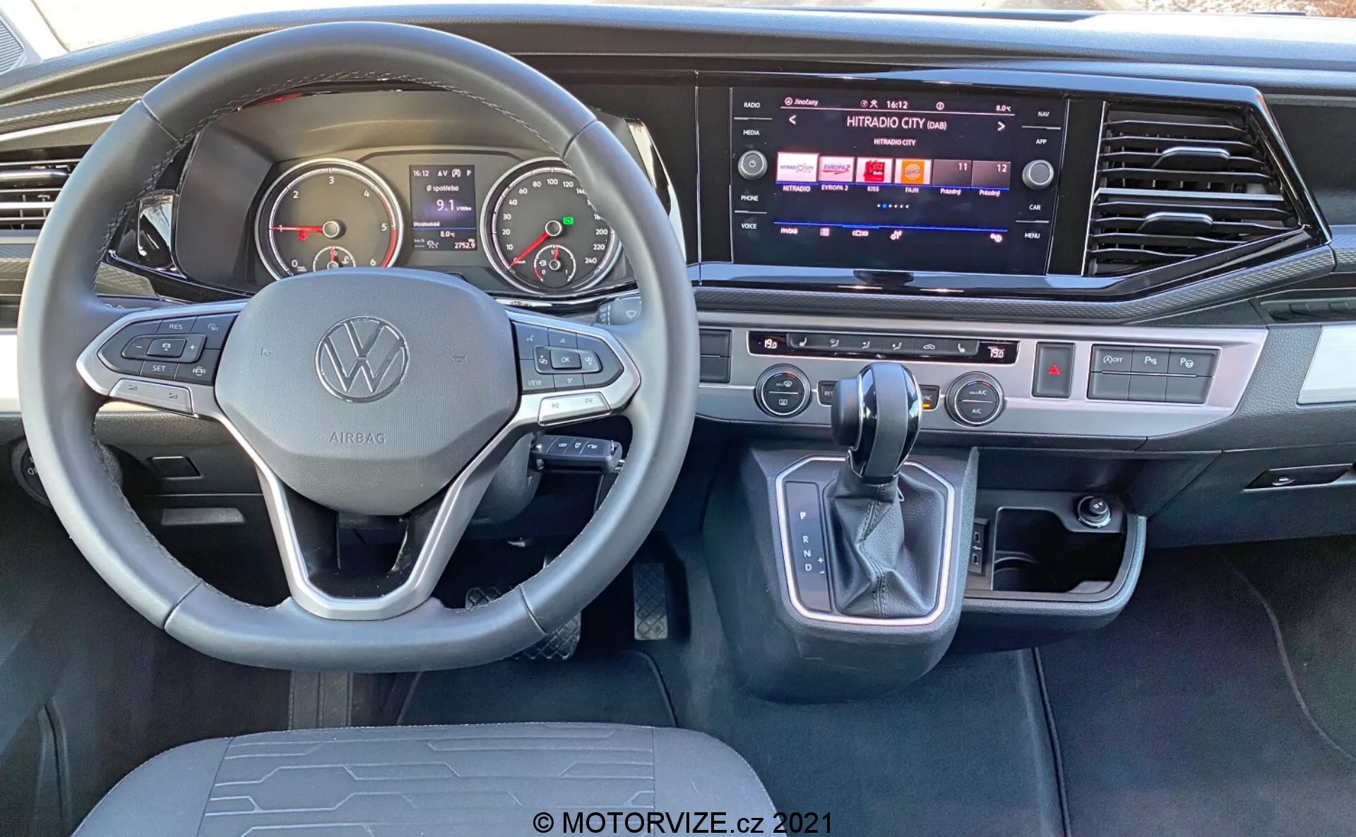 Pohľad do interiéru vozidla VW Nutzfahrzeuge Transporter 2019 T6.1 Multivan z pohľadu vodiča. Obsahuje volant s logom Volkswagen a multifunkčnými tlačidlami, analógové ukazovatele, digitálny displej, stredovú konzolu s aktívnym dotykovým infotainment systémom, dvojzónovú klimatizáciu, radiacu páku s elektronickým parkovacím tlačidlom a modernými kartáčovanými kovovými akcentmi.