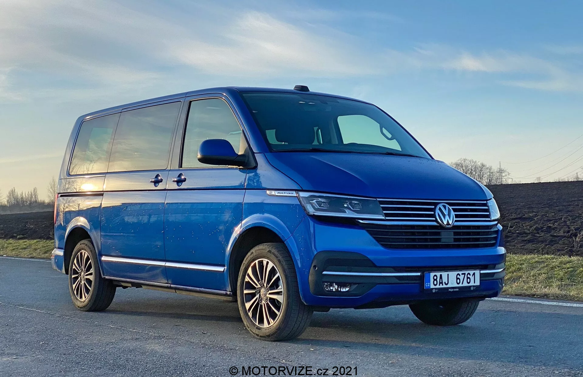 Nízky uhol pohľadu na modrý Volkswagen T6-1 Multivan 2019, ukazujúci predný ľavý kvadrant z pohľadu z troch štvrtín. Dodávka je zaparkovaná na miernom svahu cesty, s výraznými chrómovanými detailmi na mriežke chladiča, centrálnym znakom VW a modernými jednotkami svetlometov, ktoré môžu mať LED technológiu. Hmlové svetlá sú integrované do nárazníka, ktorý má aj prívody vzduchu. Bočný profil odhaľuje posuvné dvere, charakterovú líniu, ktorá sa tiahne od predného po zadný blatník, a robustné bočné zrkadlá s indikátormi smeru. Viditeľné sú aj zliatinové kolesá s viac lúčovým dizajnom a dobre udržiavaný dezén pneumatík. Obloha v pozadí ukazuje náznak západu slnka.