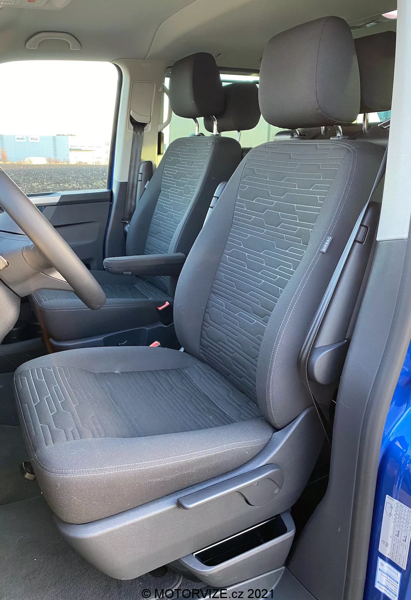 Interiér modelu VW Nutzfahrzeuge Transporter 2019 T6.1 Multivan, zobrazujúci predné sedadlo spolujazdca a časť sedadla vodiča z pohľadu zvonka na strane spolujazdca. Záber z mierne nízkeho uhla zdôrazňuje textúrovanú látku s geometrickými vzormi na sedadlách, páku manuálneho nastavenia, bezpečnostné pásy a časť stredovej konzoly a volantu. Prirodzené denné svetlo osvetľuje interiér vozidla.
