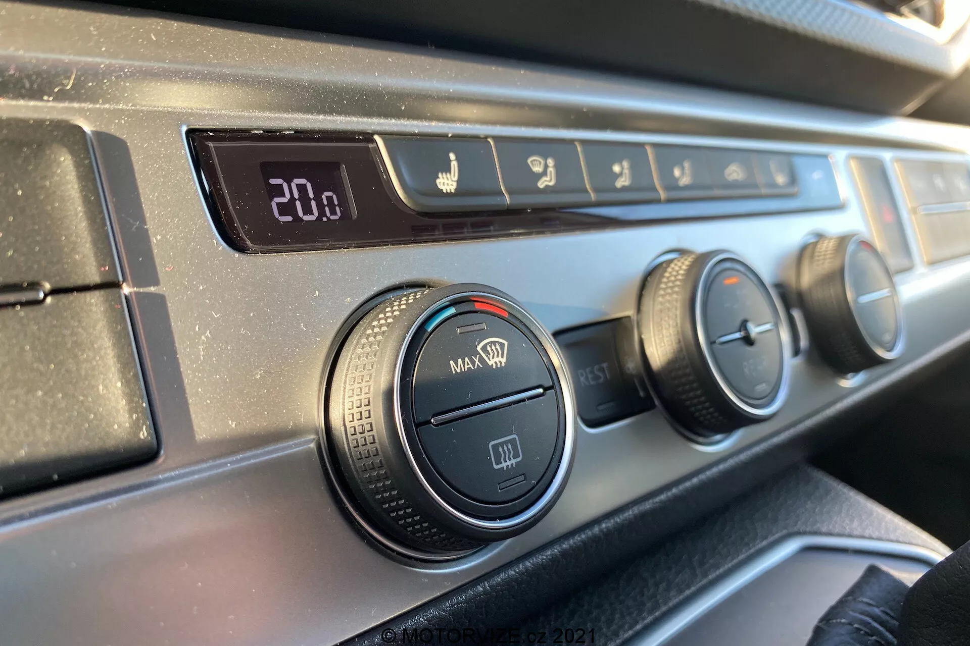Detailný pohľad na ovládací panel klimatizácie na palubnej doske vozidla VW Transporter T6.1 Multivan z roku 2019 z pohľadu spolujazdca, s digitálnym displejom teploty nastaveným na 20,0 stupňov Celzia, ovládaním smeru prúdenia vzduchu, tromi otočnými ovládačmi pre nastavenie klimatizácie a ventilátoru a tlačidlom recirkulácie, všetko s textúrovaným čiernym plastovým okrajom a hladkým tmavým povrchom palubnej dosky.