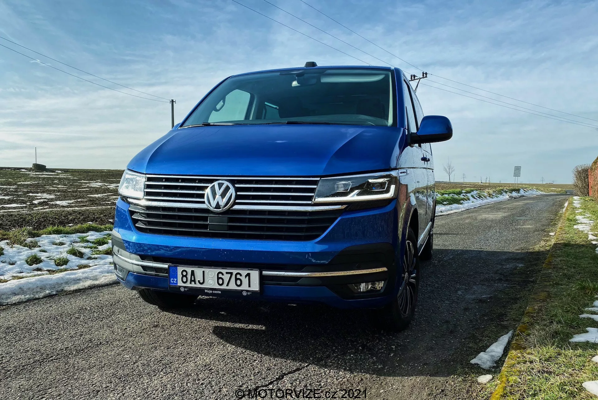 Nízky pohľad s prednou trojštvrťovou perspektívou na Volkswagen T6.1 Multivan 2019, predstavujúci prednú mriežku so horizontálnymi čiarami, výrazné VW logo v strede mriežky, svetlomety pravdepodobne s LED dennémi svetlami a bi-xenónovými alebo LED svetlami a predný nárazník s hmlovými svetlami a prívodmi vzduchu. Obrázok zobrazuje časť ľavej strany vrátane dverí vodiča a blatníka, pričom viditeľná je aj kapota a čelné sklo. Auto je hlbokomodré a zaparkované na vidieckej ceste so snehom na zemi a čistou modrou oblohou nad ním.