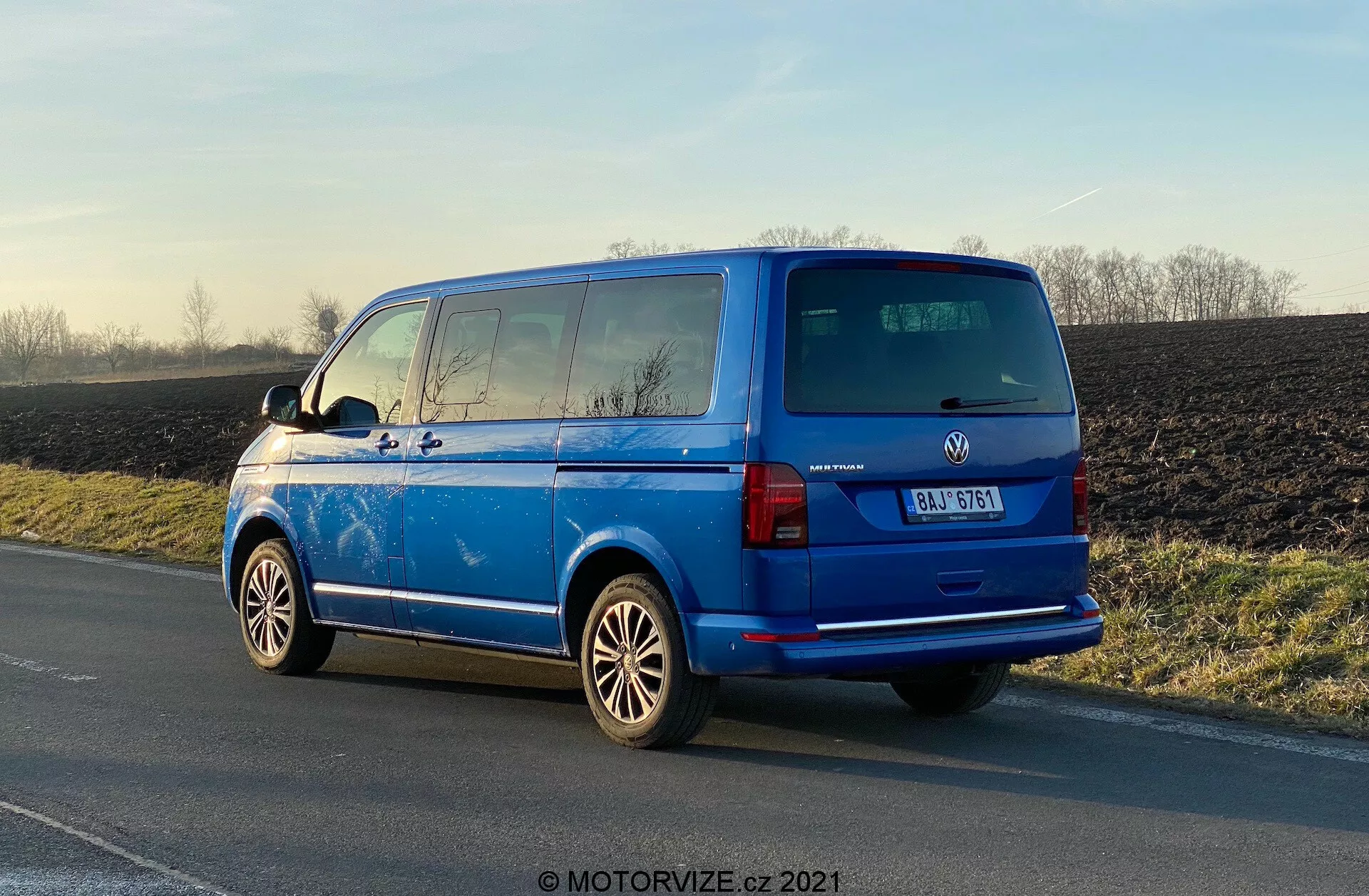 Žiarivo modrý Volkswagen Transporter 2019 (T6-1 Multivan) zaparkovaný na krajnici, zadný pohľad z troch štvrtín ukazuje zadné svetlá, zadné okno, zadné dvere, odznak výrobcu, detaily modelu; viditeľné zliatinové kolesá, vonkajšie spätné zrkadlá, vyhrievacie linky zadného okna, zatmavené okná druhého radu, strešnú anténu; zaparkovaný v prirodzenom dennom svetle so zvýrazneným lesklým povrchom, s vidieckym pozadím.