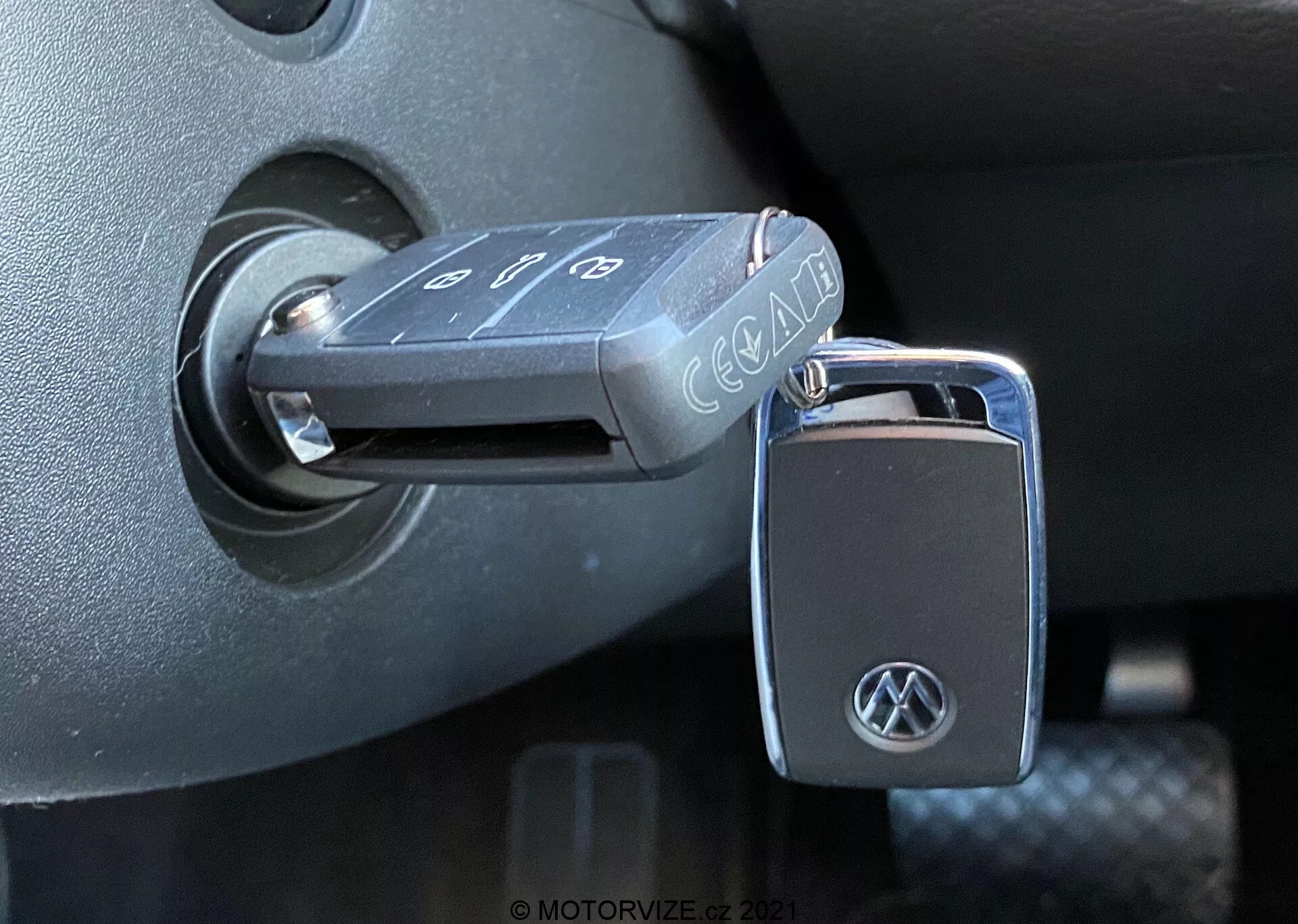 Detailný pohľad na sklopný kľúč Volkswagen vložený do zapaľovania vozidla VW Nutzfahrzeuge T6-1 Multivan z roku 2019. Kľúč s tlačidlami na zamknutie, odomknutie a alarm má logo VW na kľúčenke. Zapaľovanie sa nachádza v tmavom plastovom prostredí stĺpika riadenia, čo naznačuje, že zapaľovanie je v polohe 'vypnuté'. Fotografia, urobená z bočného uhla pri dennom svetle, zvýrazňuje detailné textúry a povrchy interiéru vozidla.