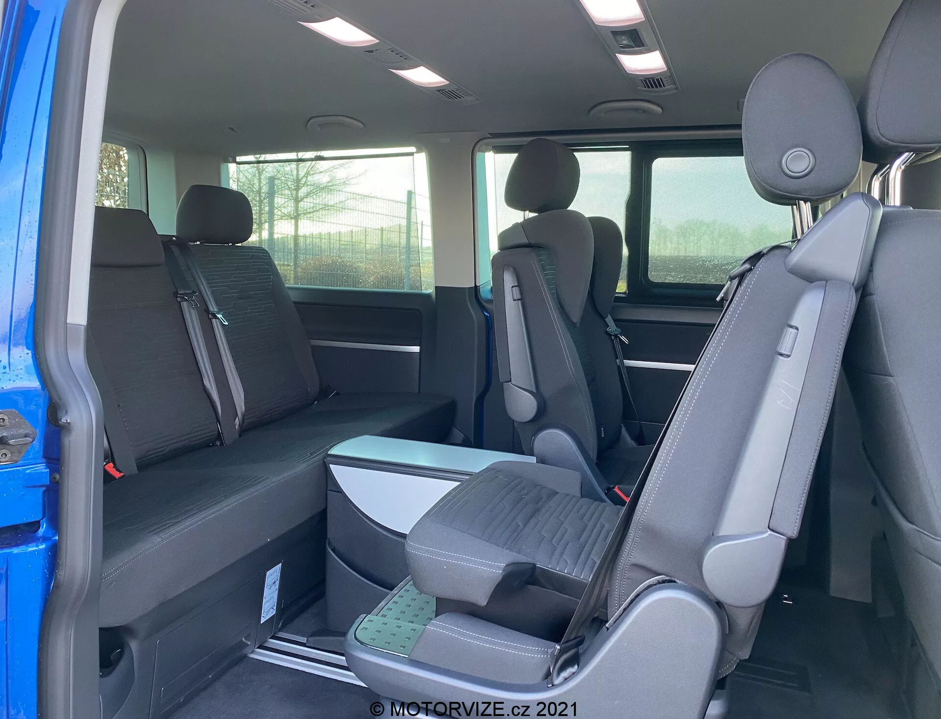 Interiér VW Transporter T6.1 Multivan 2019 zobrazujúci druhý a tretí rad sedadiel z pohľadu zadnej cestujúcej strany s konfiguráciou sedadiel 2-3-2 a tmavým látkovým čalúnením. Čiastočný pohľad na exteriér cez otvorené dvere, opierky hlavy na všetkých sedadlách, zvýšený tretí rad sedadiel pre lepšiu viditeľnosť.