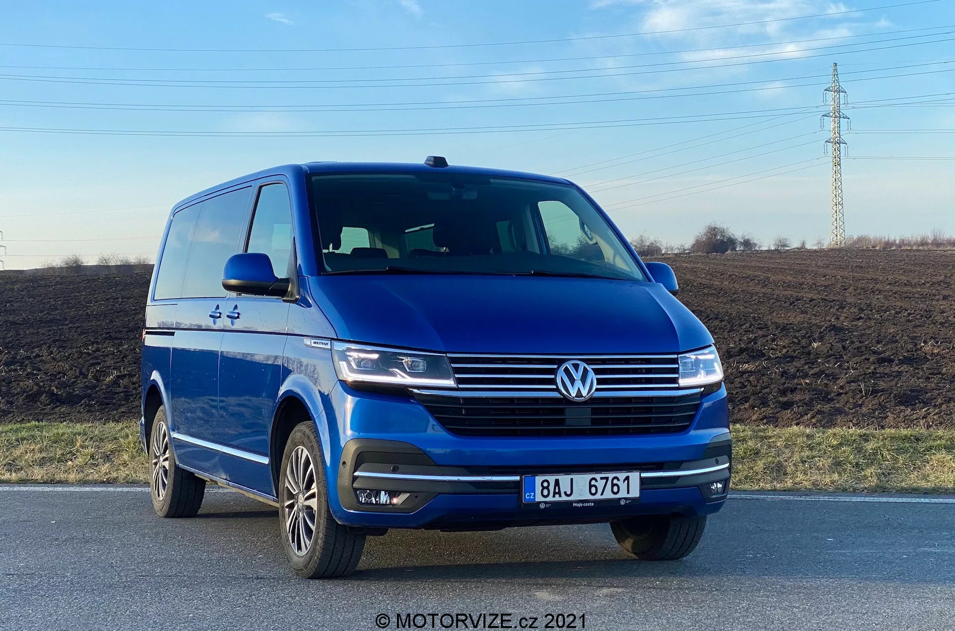 Obrázok zobrazuje modrý Volkswagen VW-Nutzfahrzeuge Transporter 2019 (T6-1 Multivan) zachytený z nízkeho frontálneho uhla, zobrazujúci čelný ľavý štvrťový pohľad s jasným výhľadom na prednú časť a profil vodičovej strany. Vozidlo je na ceste, zvýrazňujúce prvky ako prominentný znak Volkswagen, elegantné svetlomety, tvarované línie kapoty, integrované hmlové svetlá na nárazníku a viaclúčové zliatinové kolesá. Fotografia urobená počas denného svetla zdôrazňuje vonkajší dizajn vozidla a technológie asistencie vodiča, ako sú parkovacie senzory.